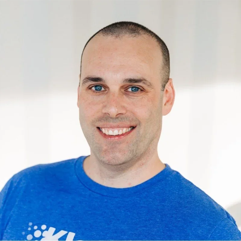 A smiling man with blue eyes and short hair, wearing a blue T-shirt, standing indoors against a neutral background.