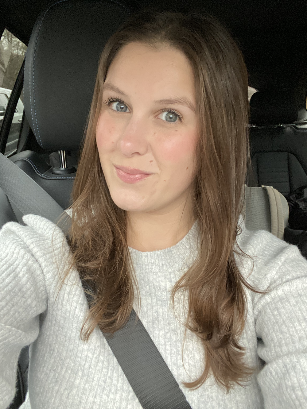 A woman sitting in a car wearing a seatbelt, with long brown hair, blue eyes, and a light-colored sweater, taking a selfie.