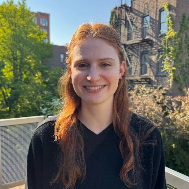 A young woman with long red hair smiling, standing outdoors on a balcony with trees and buildings in the background on a sunny day.