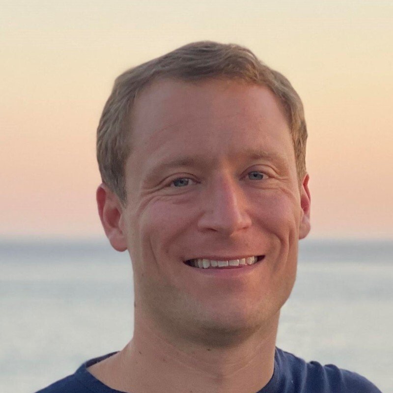 A man with short, light brown hair smiling at the camera outdoors near the water at sunset.