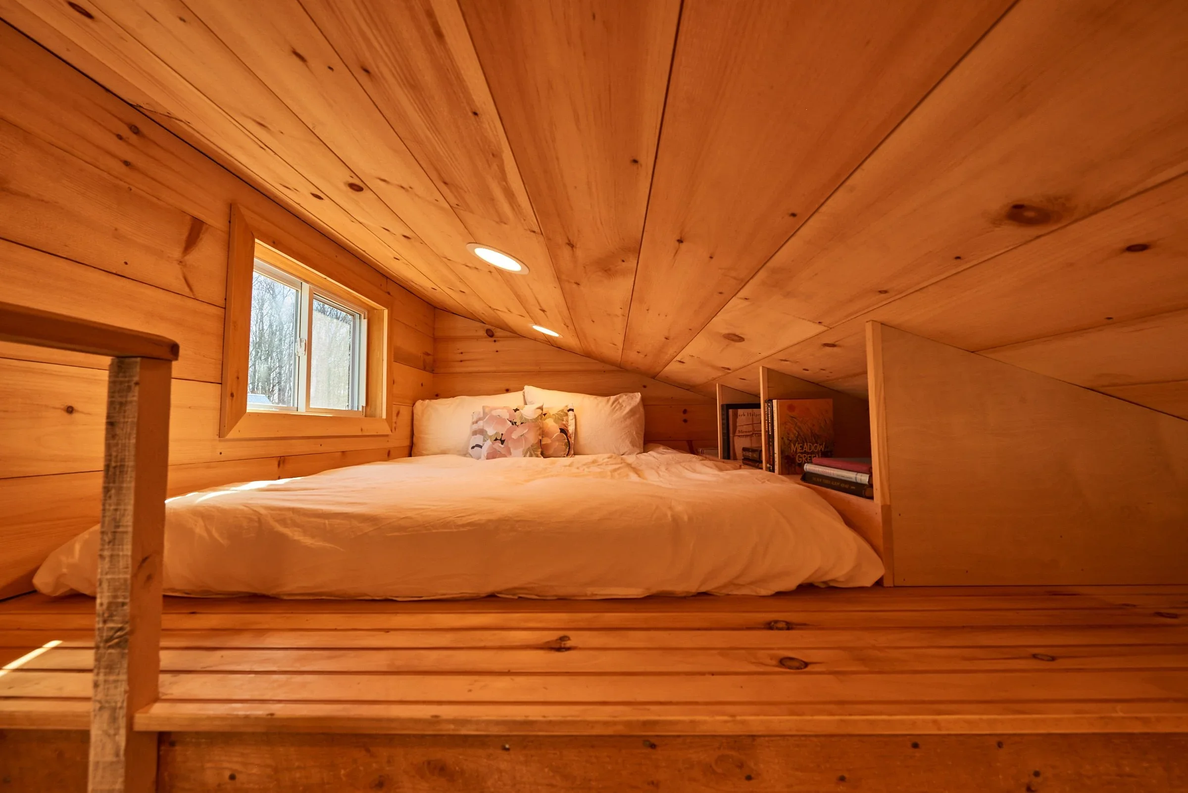 Wooden bedroom with slanted ceiling, small window, bed with white bedding and pillows, built-in bookshelf on right side.