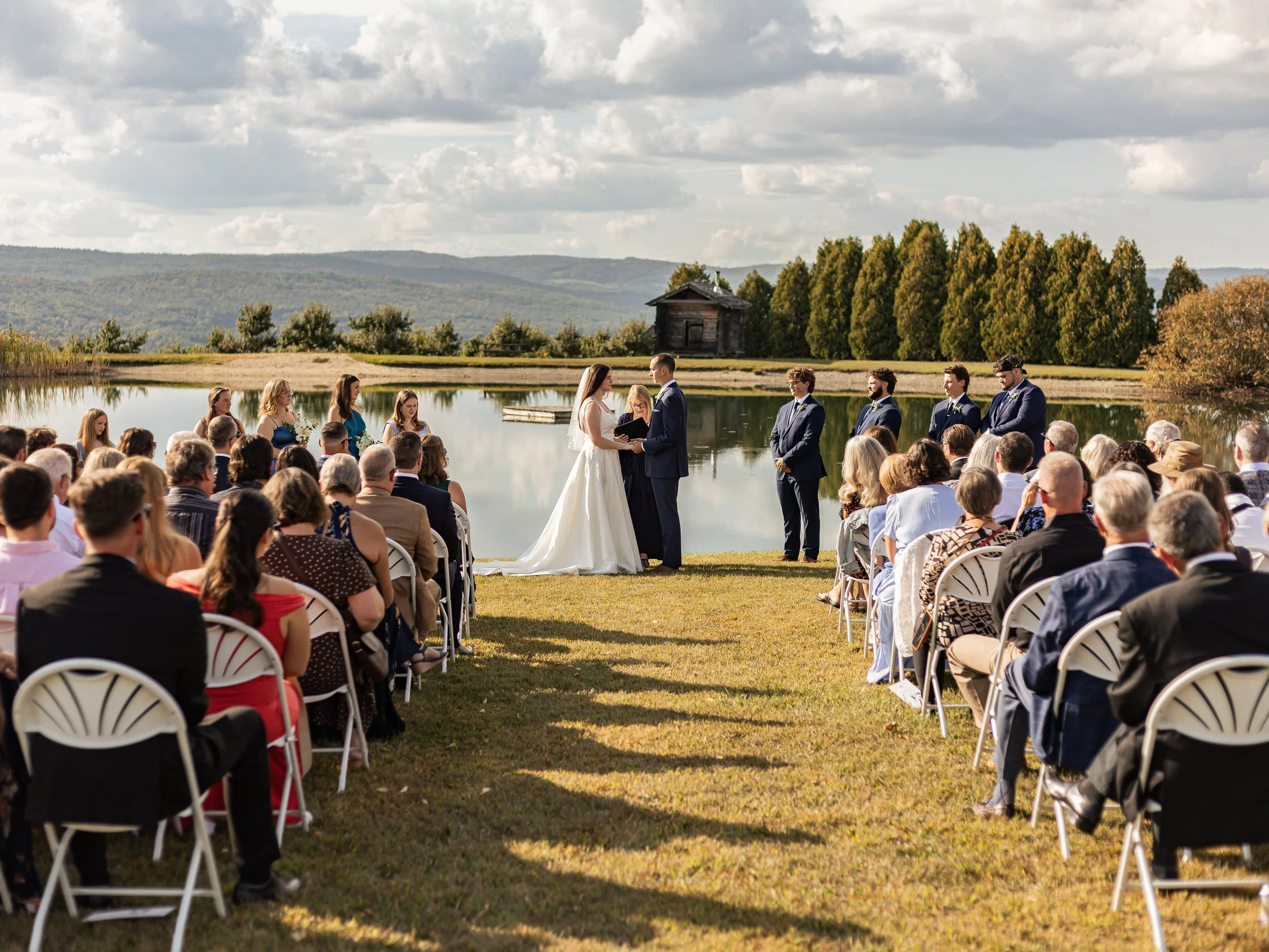 A wedding ceremony taking place outdoors by a lake, with the bride and groom exchanging vows surrounded by guests, a scenic landscape of mountains, trees, and a cloudy sky in the background.