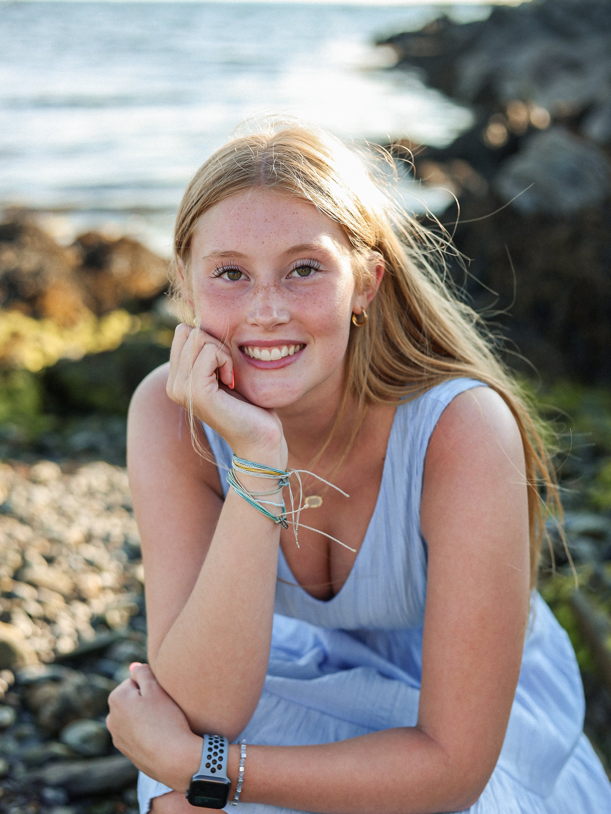 A young woman with long blonde hair, wearing a light blue dress, sitting outdoors by the rocky shoreline, smiling at the camera with her left hand resting on her chin.