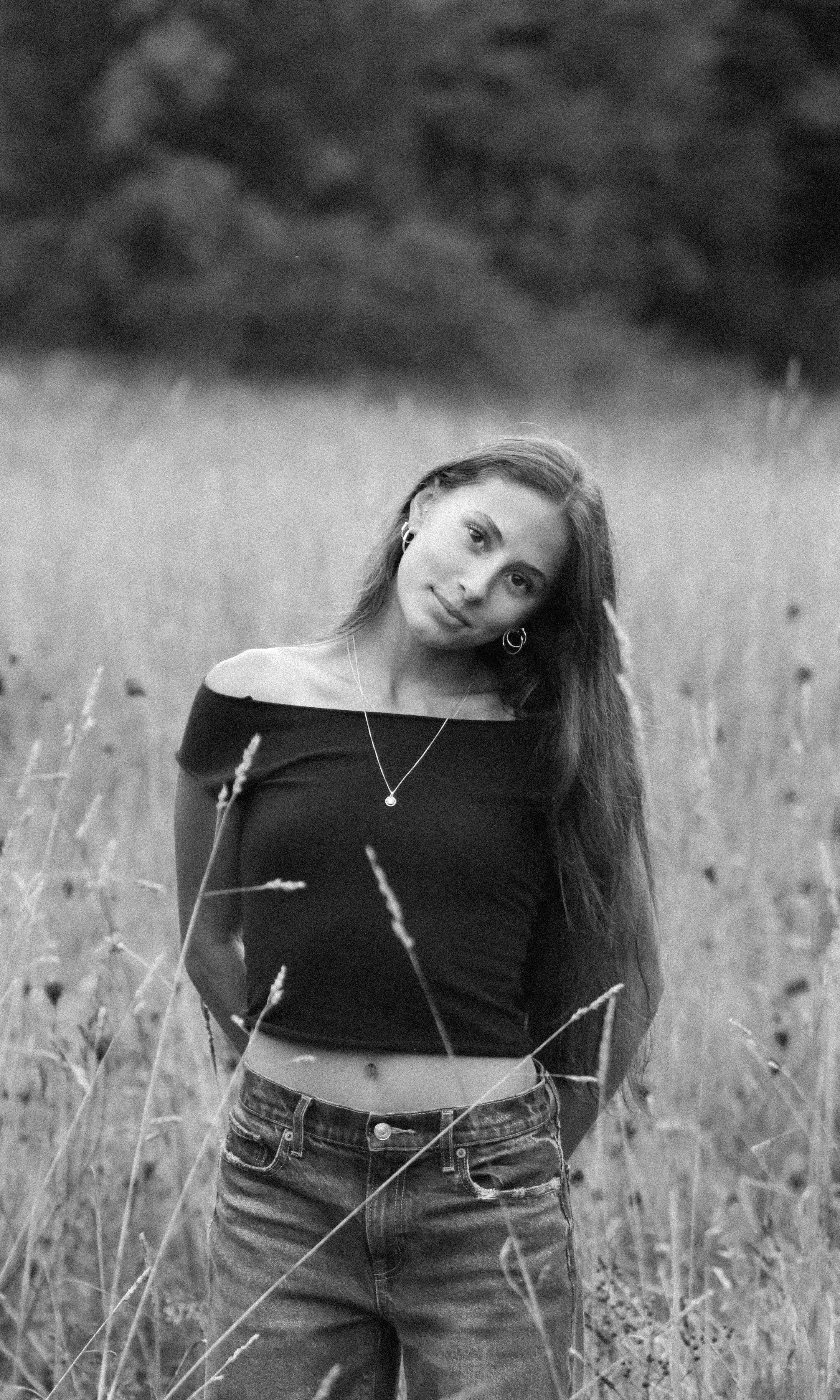 A young woman in a black off-shoulder top and jeans standing in a field of tall grass, looking at the camera with a slight tilt of her head.