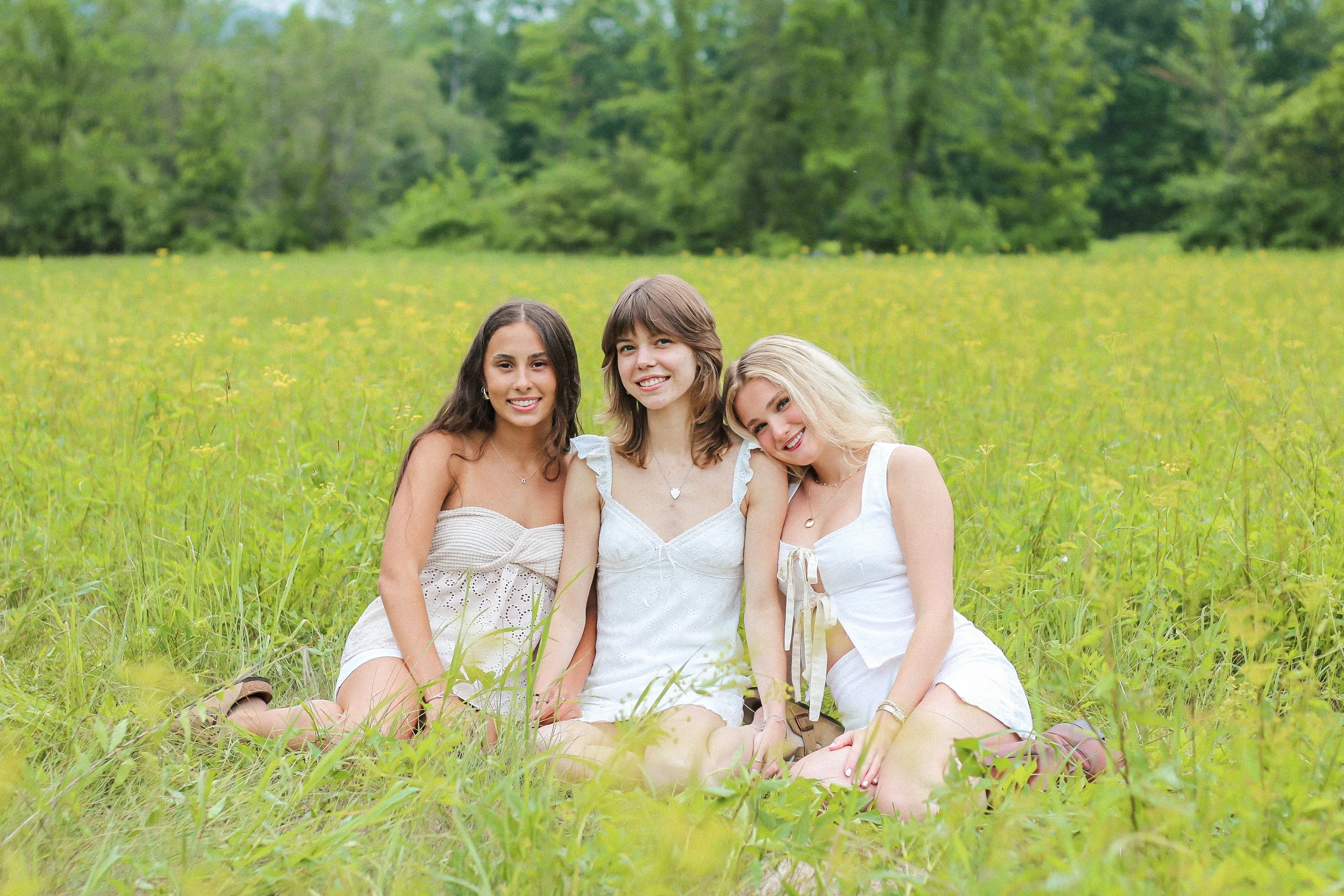 Three young women sitting on grass in a field, smiling, surrounded by green trees.