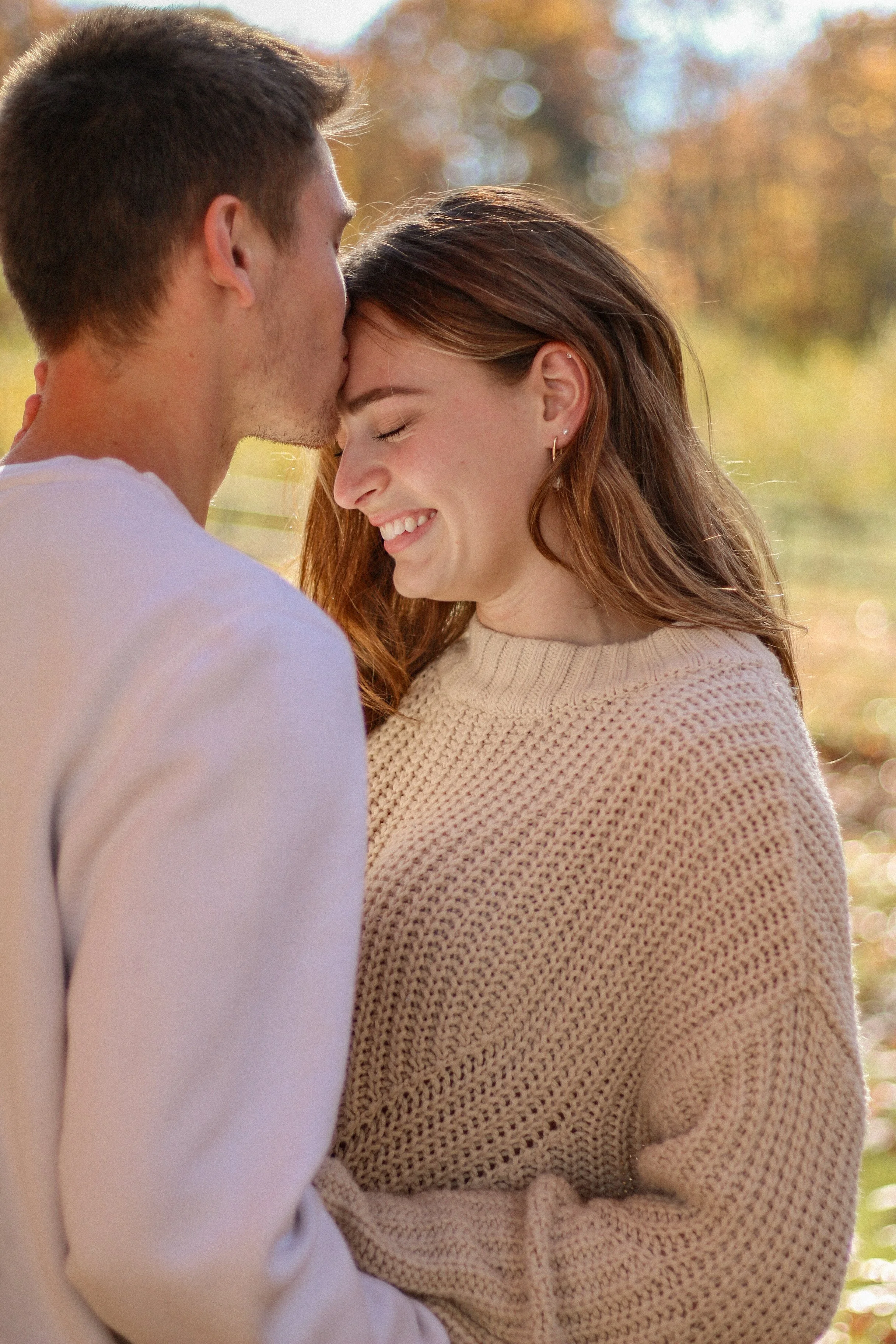 A young couple sharing an intimate moment outdoors, with the man kissing the woman's forehead and the woman smiling with her eyes closed, in a setting with fall foliage in the background.