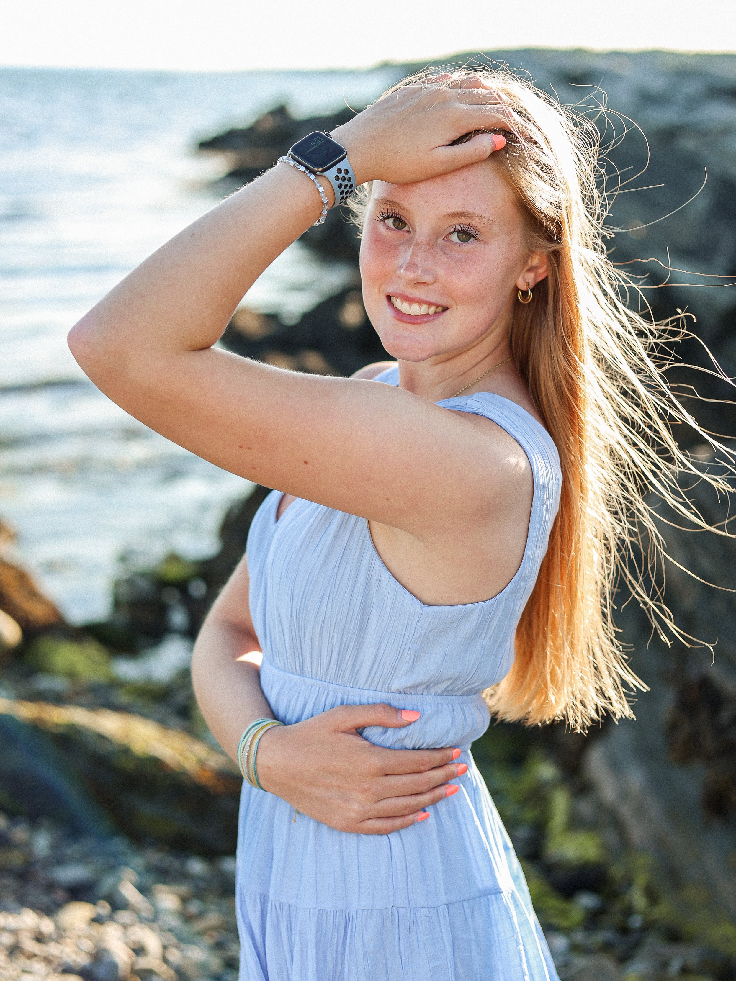 Young woman with long red hair on the beach, smiling, wearing a light blue dress, jewelry, and a smartwatch, with rocks and water in the background.