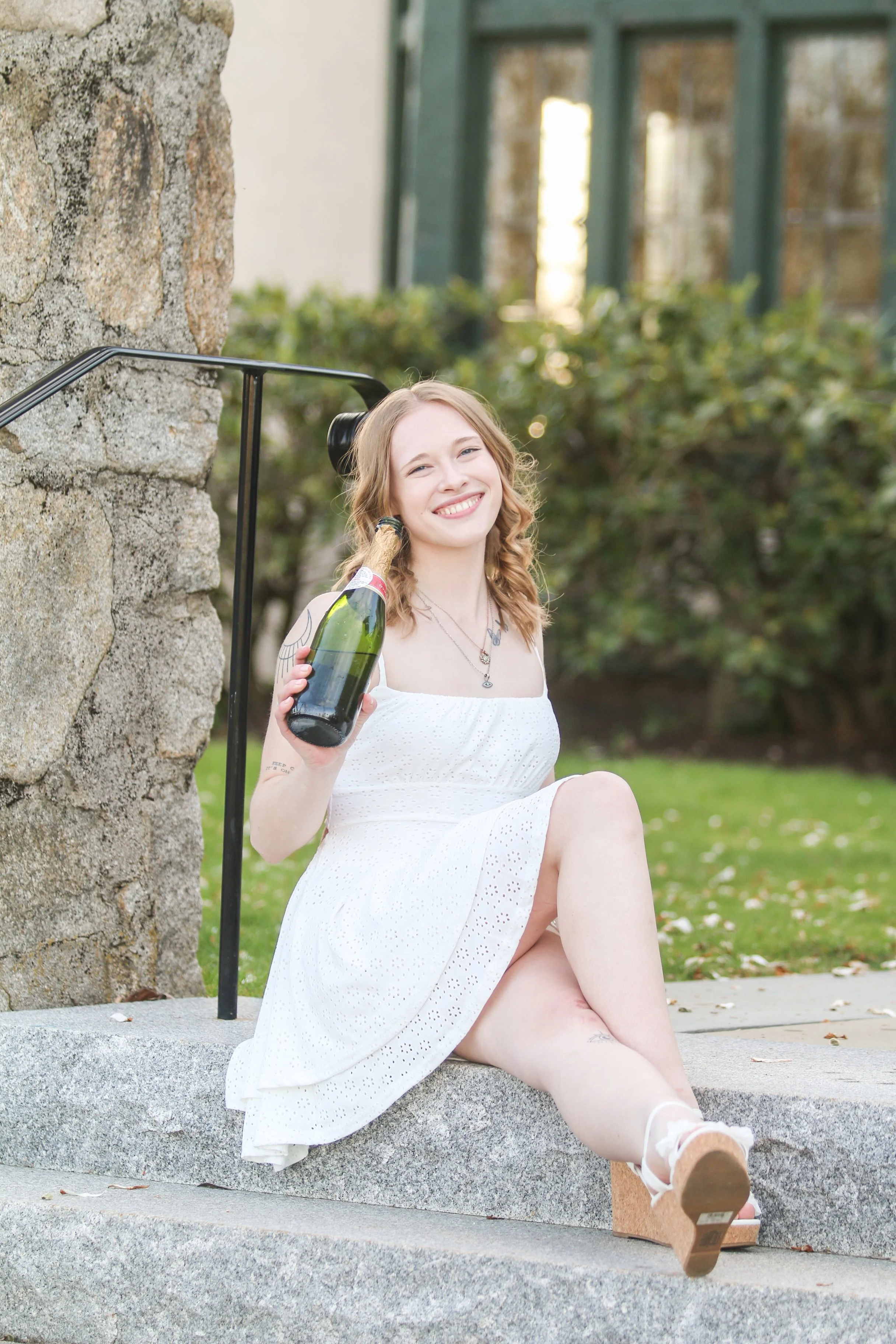 A young woman with wavy, shoulder-length hair, wearing a white dress, sitting on outdoor concrete steps near a stone wall, smiling, holding a bottle of sparkling wine, with a background of greenery and a structure with large windows.