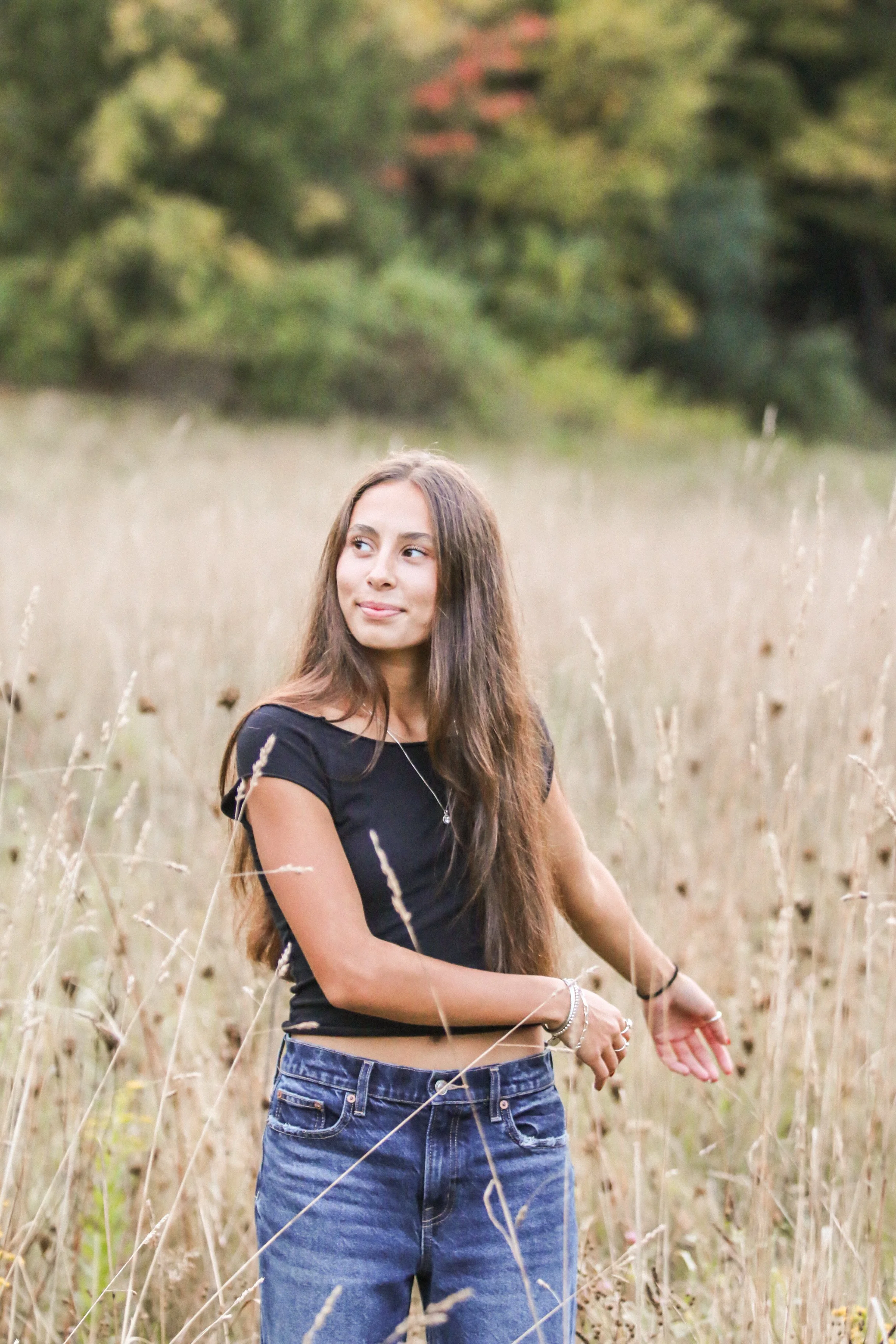 A young woman with long brown hair standing in a field of tall grass, looking to her left with a slight smile, wearing a black T-shirt and blue jeans.