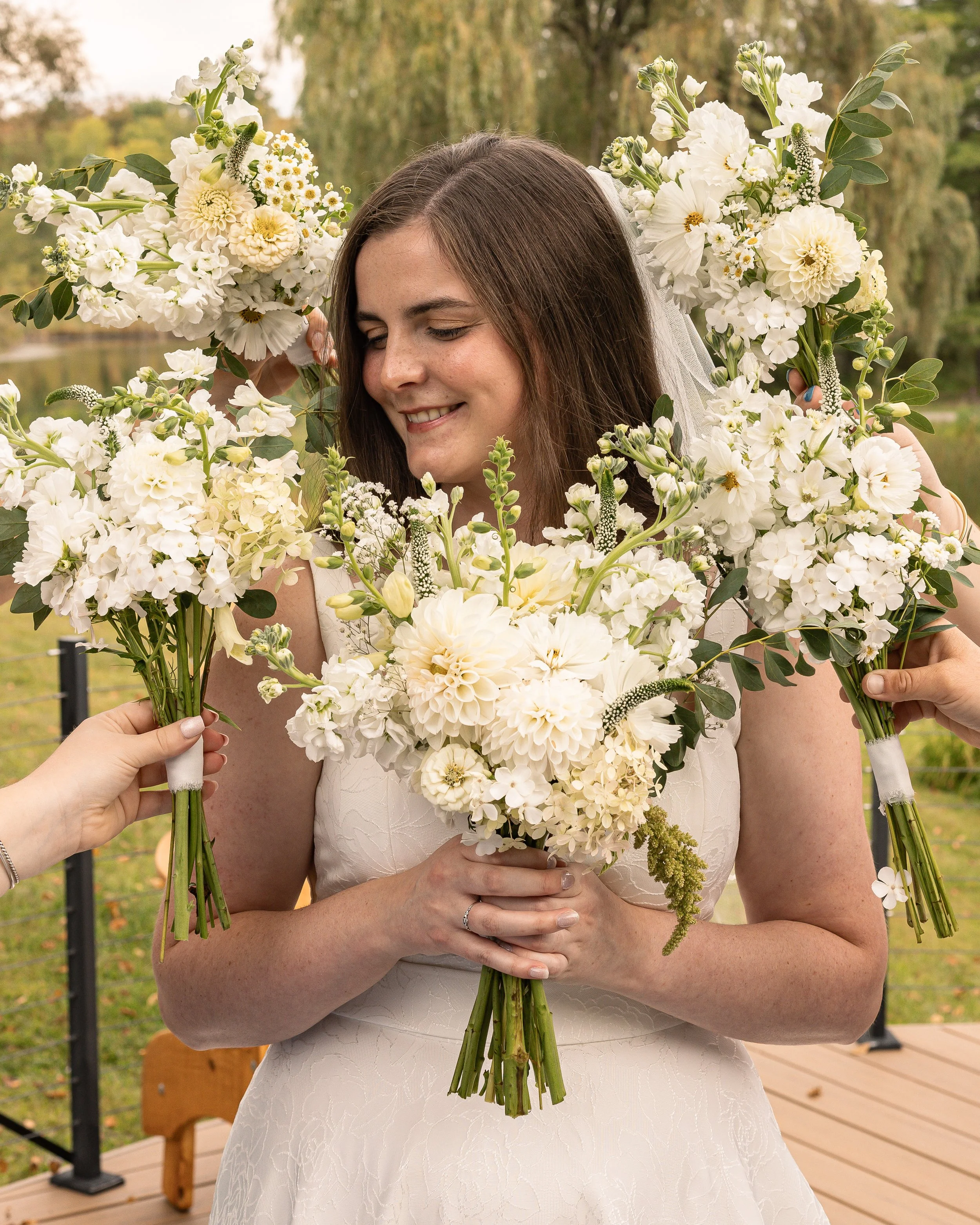 Bride holding a bouquet of white flowers with two people offering additional bouquets in a park setting.