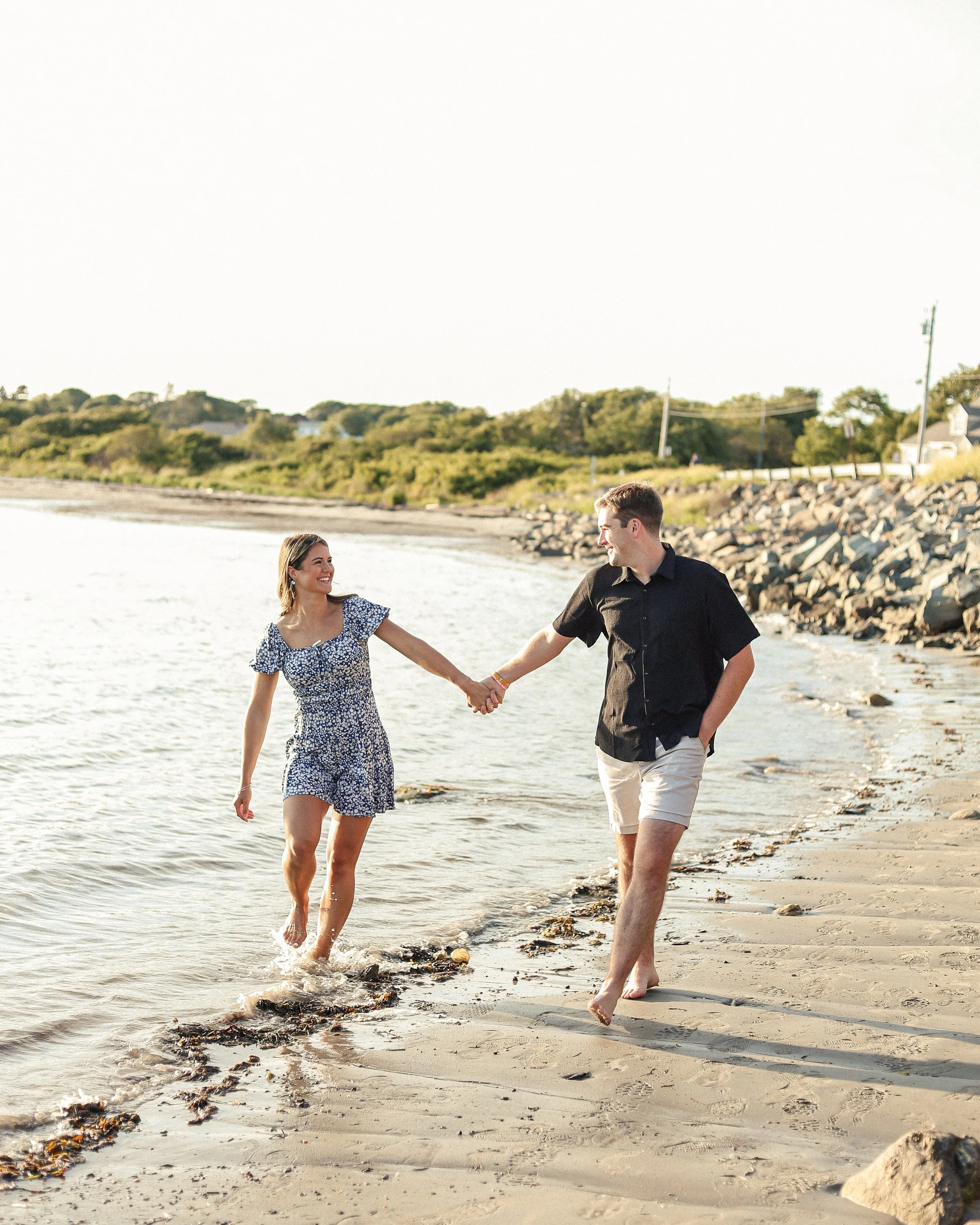 A young couple walking hand in hand along the shoreline of a beach during sunset, smiling and enjoying the moment.