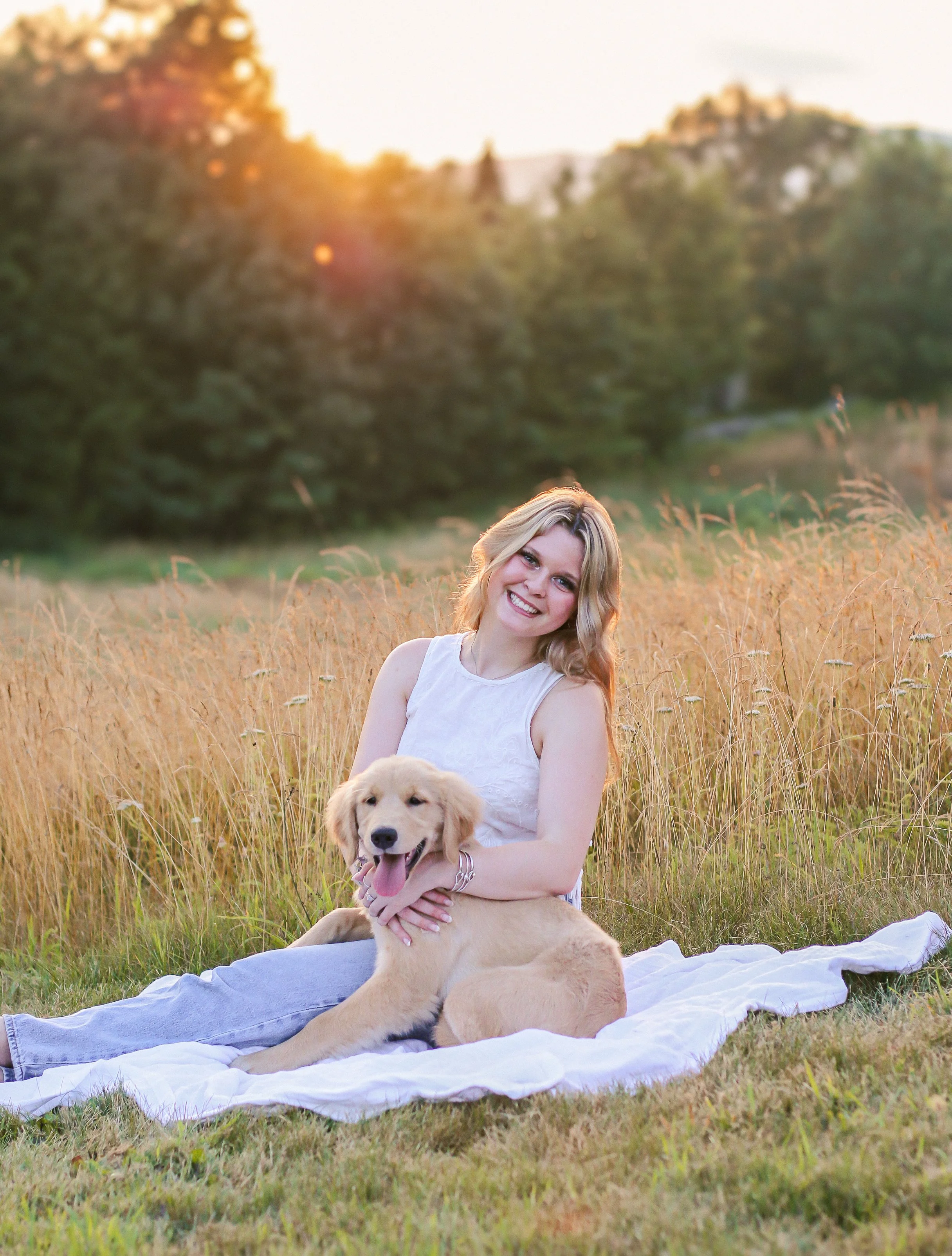 A woman in a white sleeveless top and light jeans sitting on a white blanket in a field of tall grass with a golden retriever puppy, during sunset with trees and hills in the background.