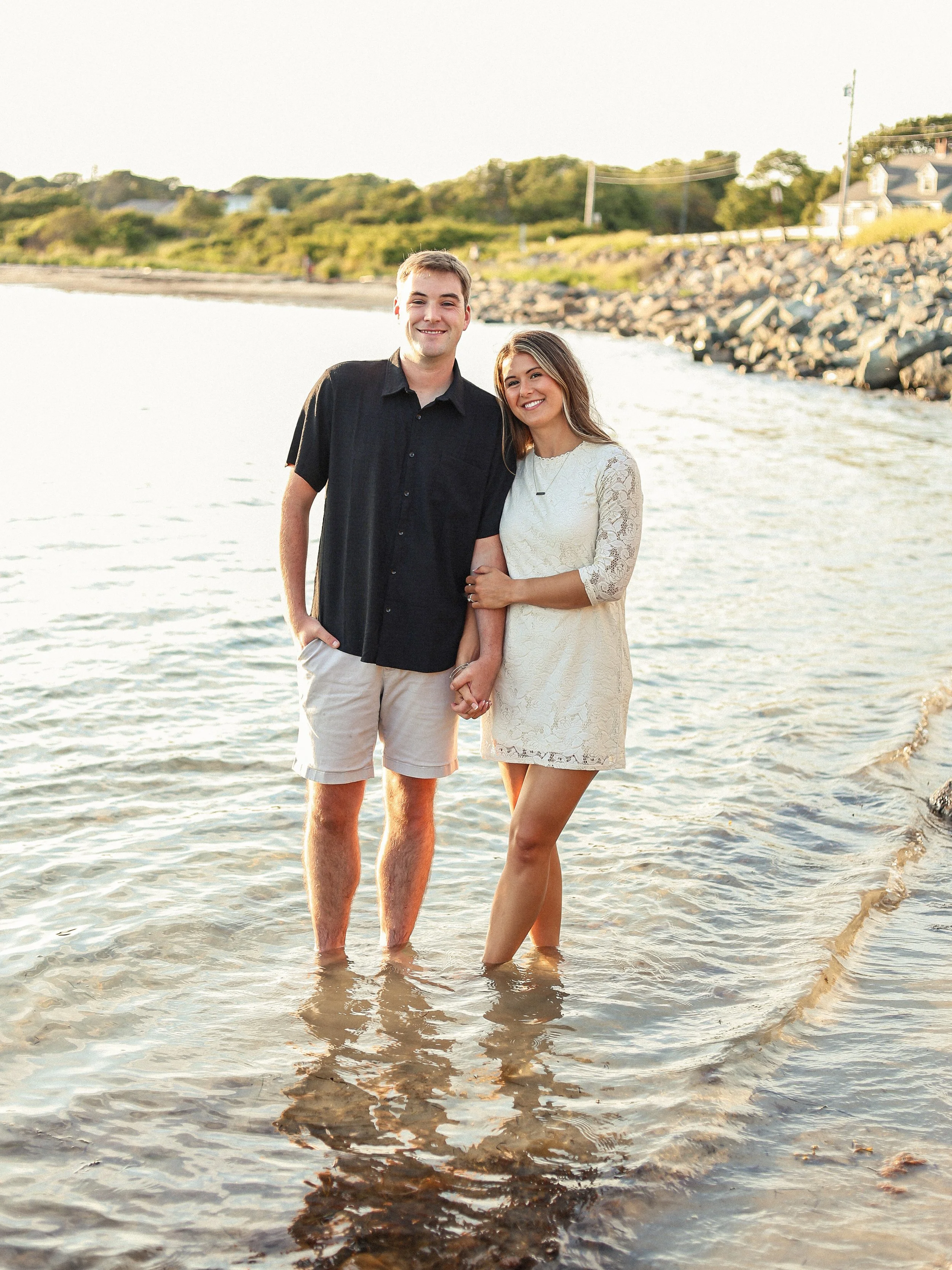 Young couple standing in shallow water at the beach, holding hands and smiling, with a rocky shoreline and houses in the background during sunset.