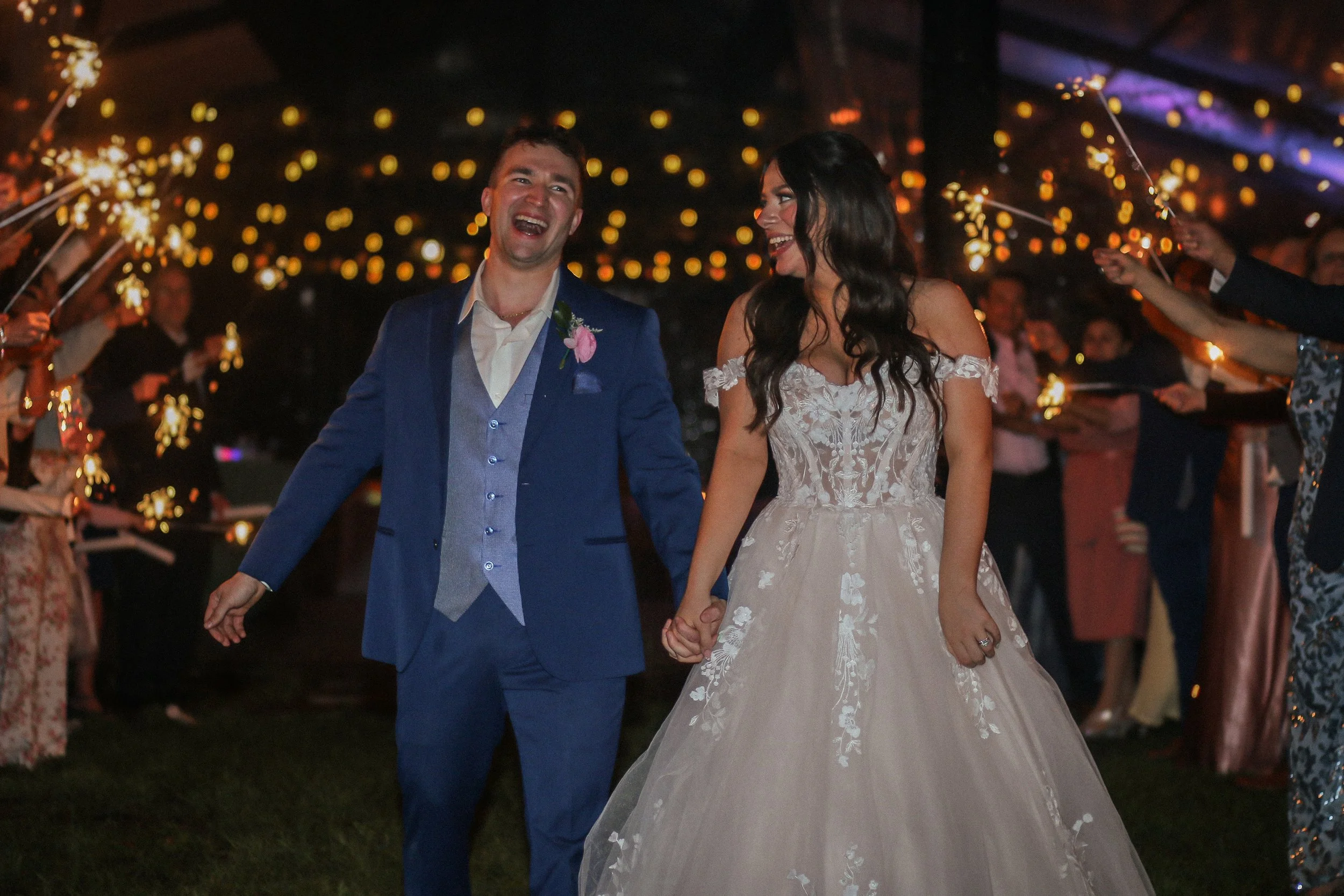 A newlywed couple holding hands and smiling at their wedding reception, surrounded by guests holding sparklers at night.