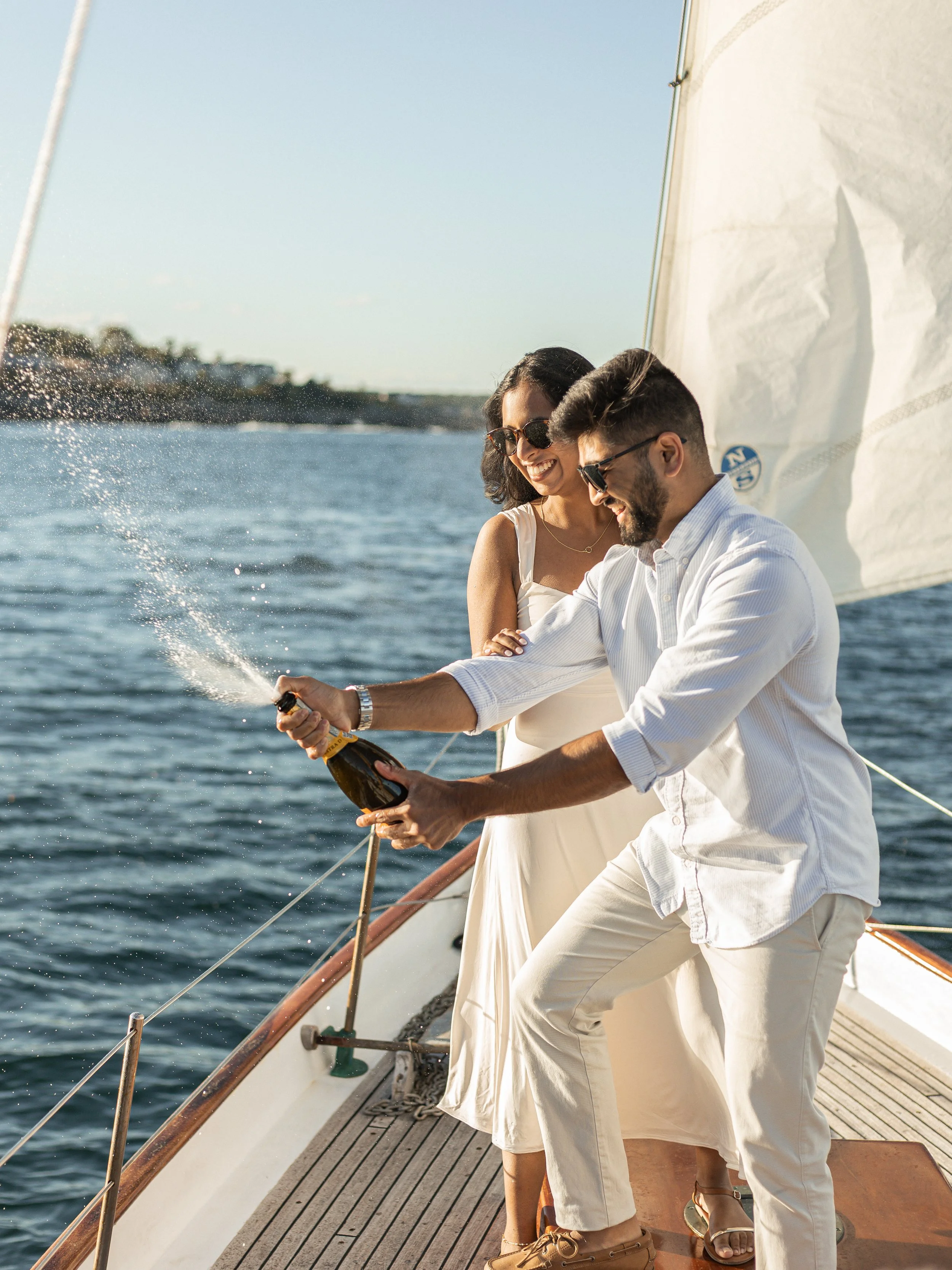 A man and woman celebrating on a sailboat, with the man opening a champagne bottle while the woman watches and smiles.