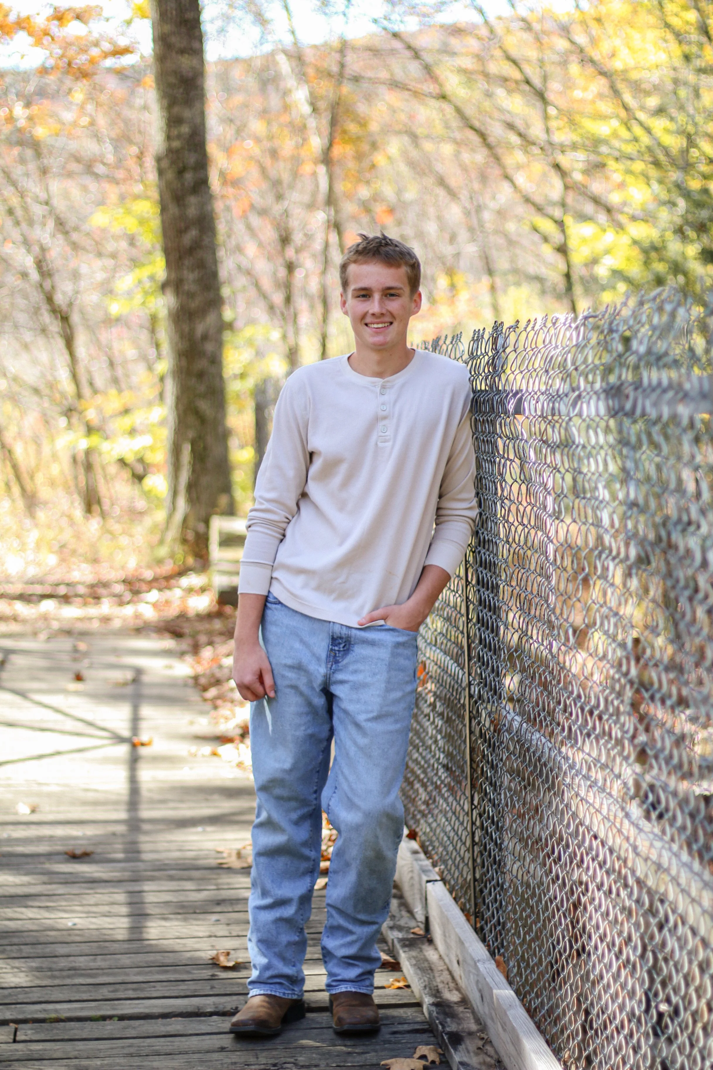 A young man leaning against a chain-link fence on a wooden pathway in a wooded area during autumn, smiling at the camera.