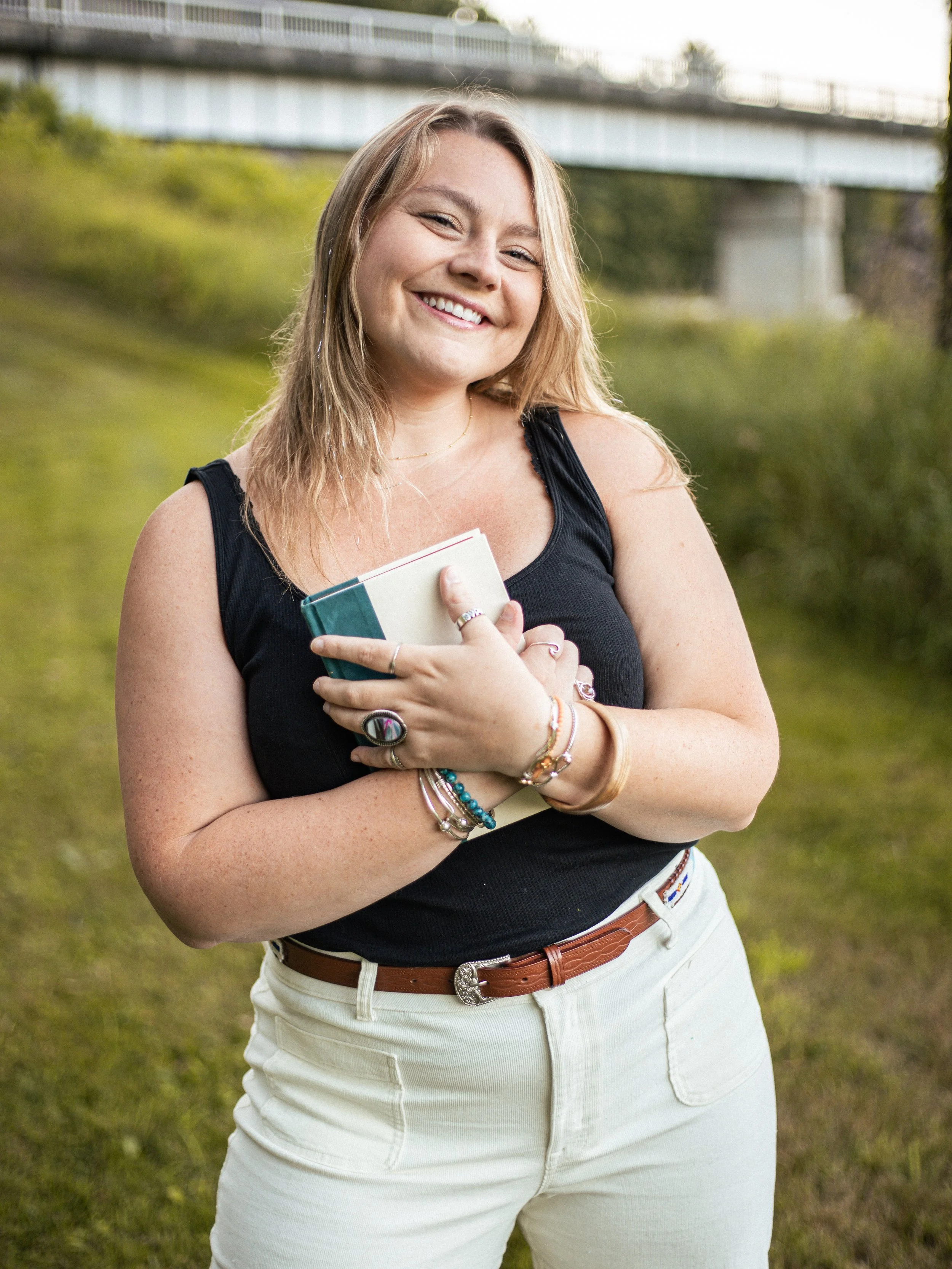 A woman with blonde hair smiling outdoors by a green grassy area, holding a book or notebook close to her chest, wearing a black sleeveless top, beige pants, and various bracelets.