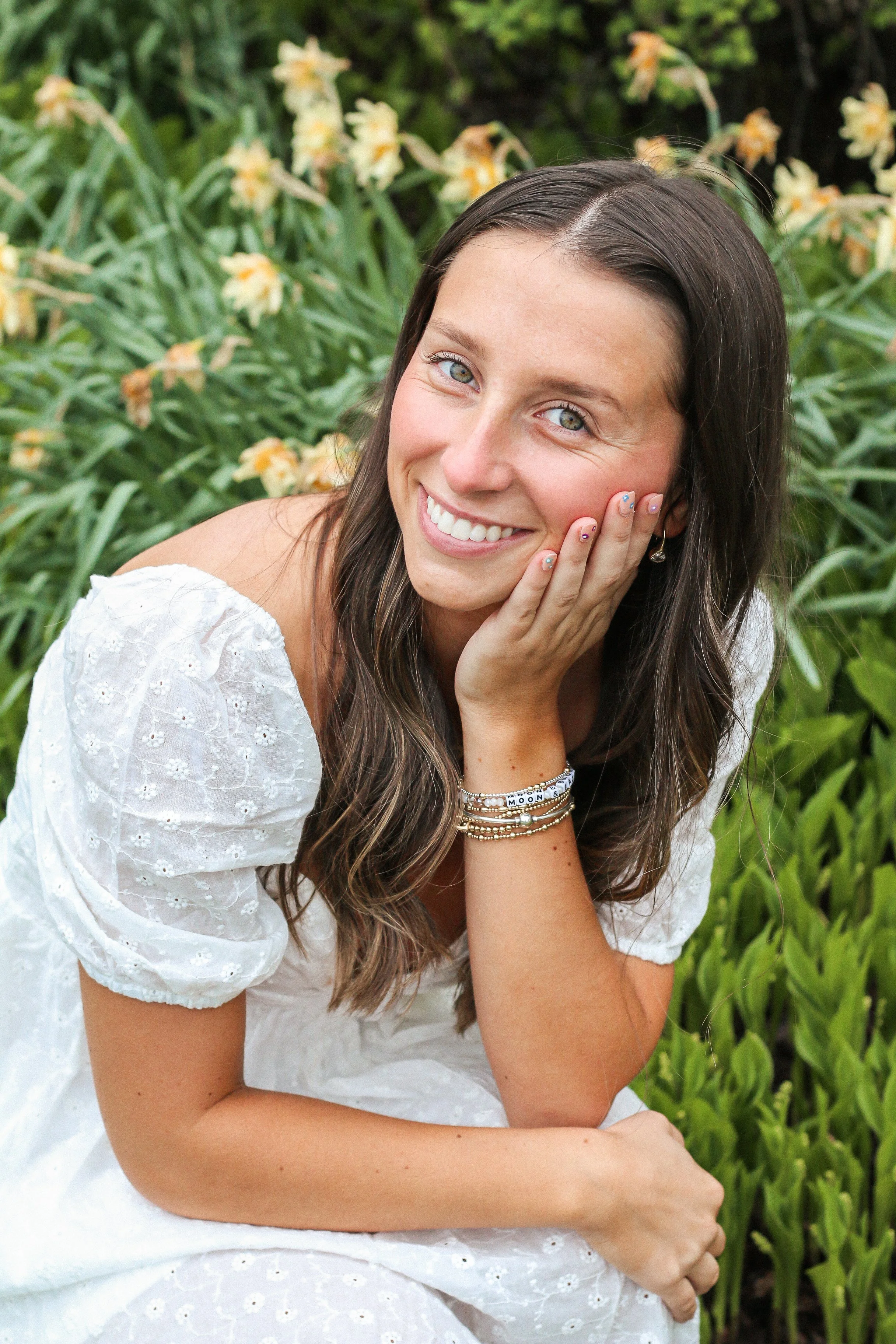 A smiling young woman with long brown hair and blue eyes, wearing a white dress with puffed sleeves, sitting outdoors among green plants and light yellow flowers.