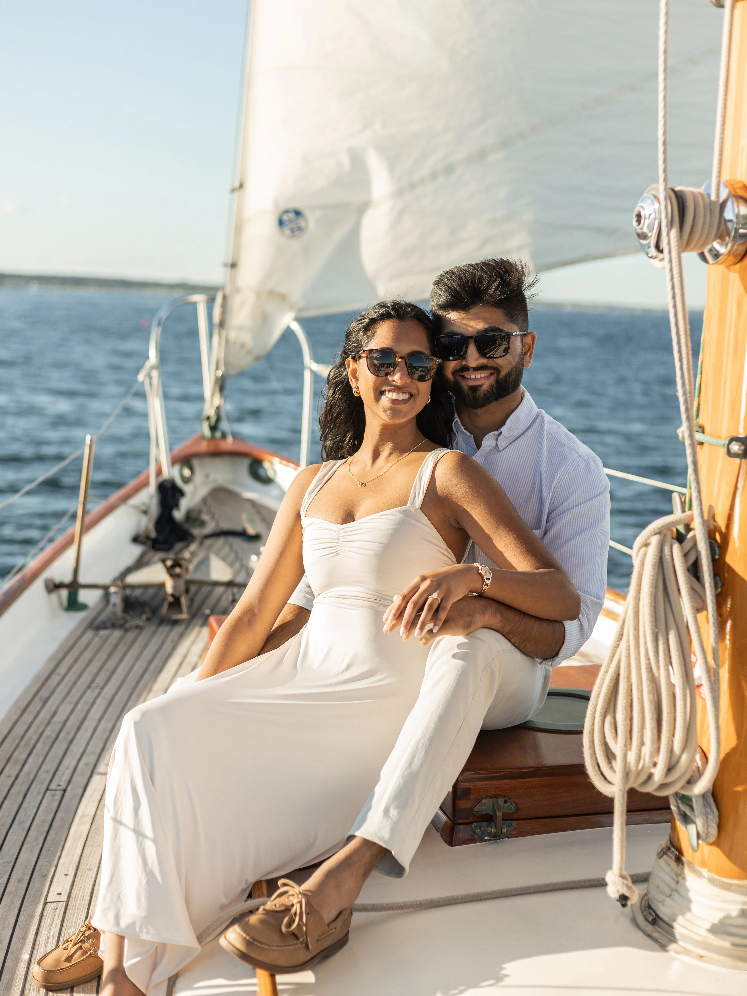 A couple wearing sunglasses sitting on a boat smiling at the camera with water and a sail in the background.