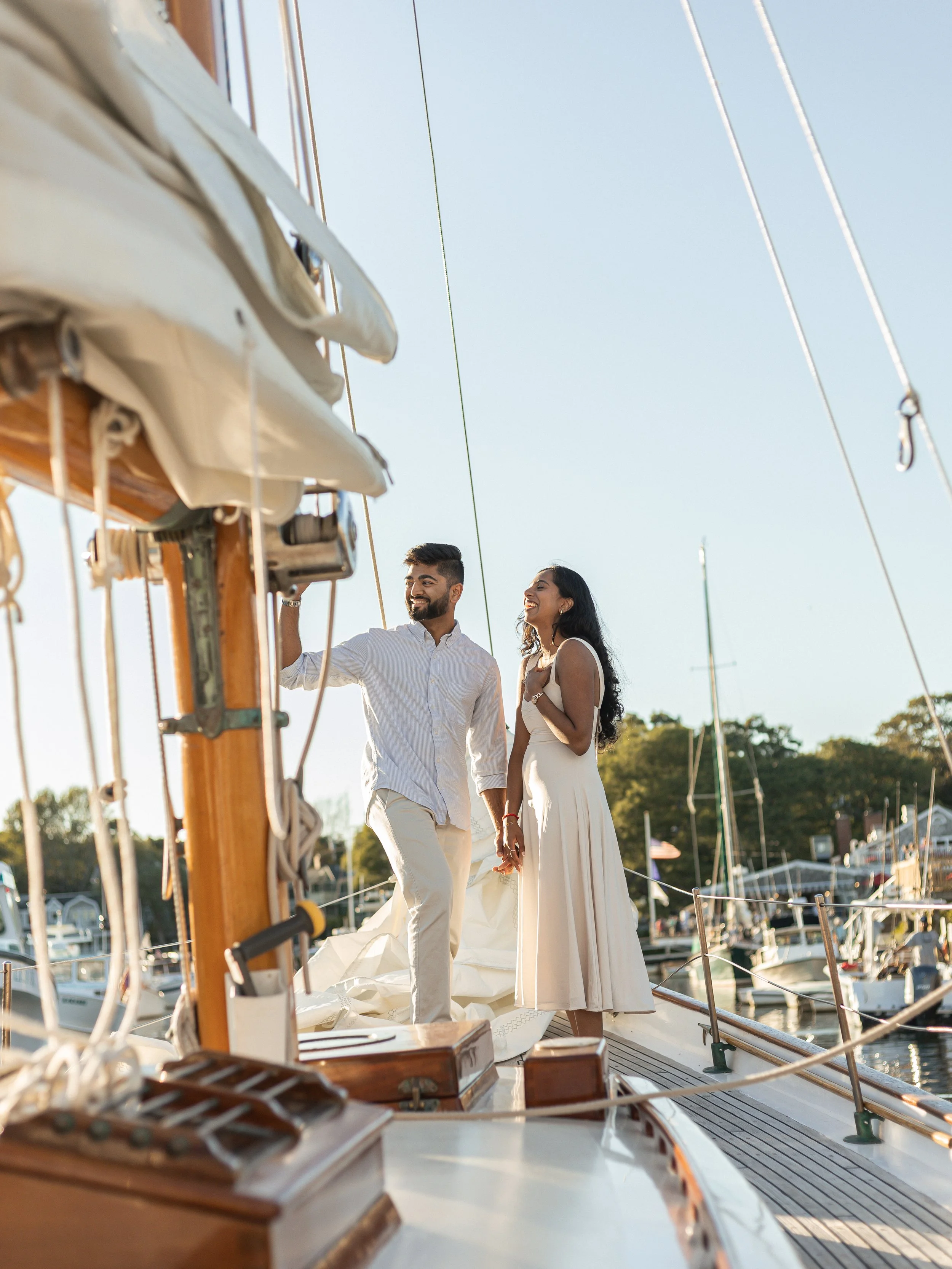 A couple holding hands on a sailboat, smiling, with a marina in the background during daytime.