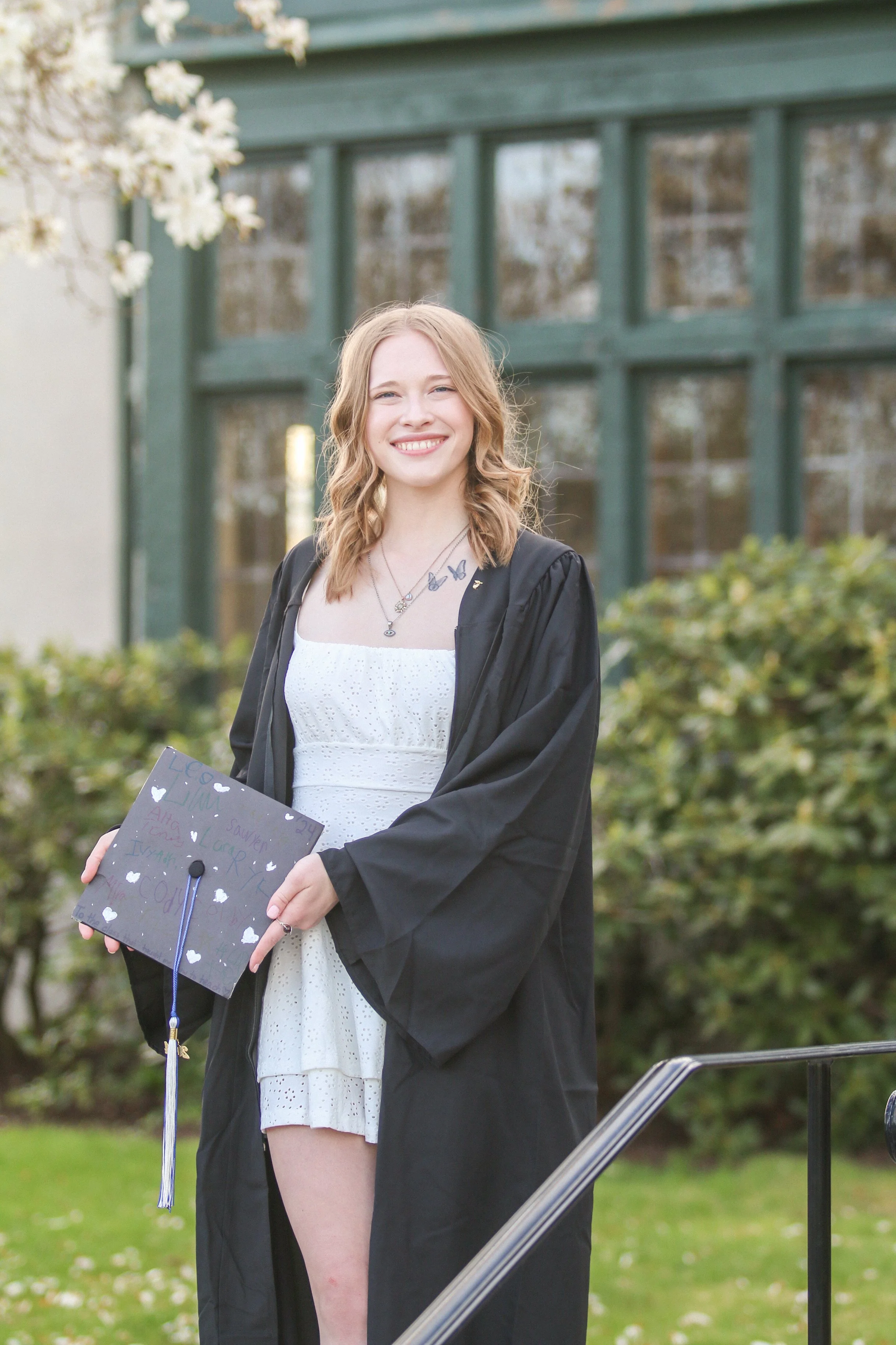 A young woman in a white dress and black graduation gown holding a decorated graduation cap outdoors, smiling at the camera.