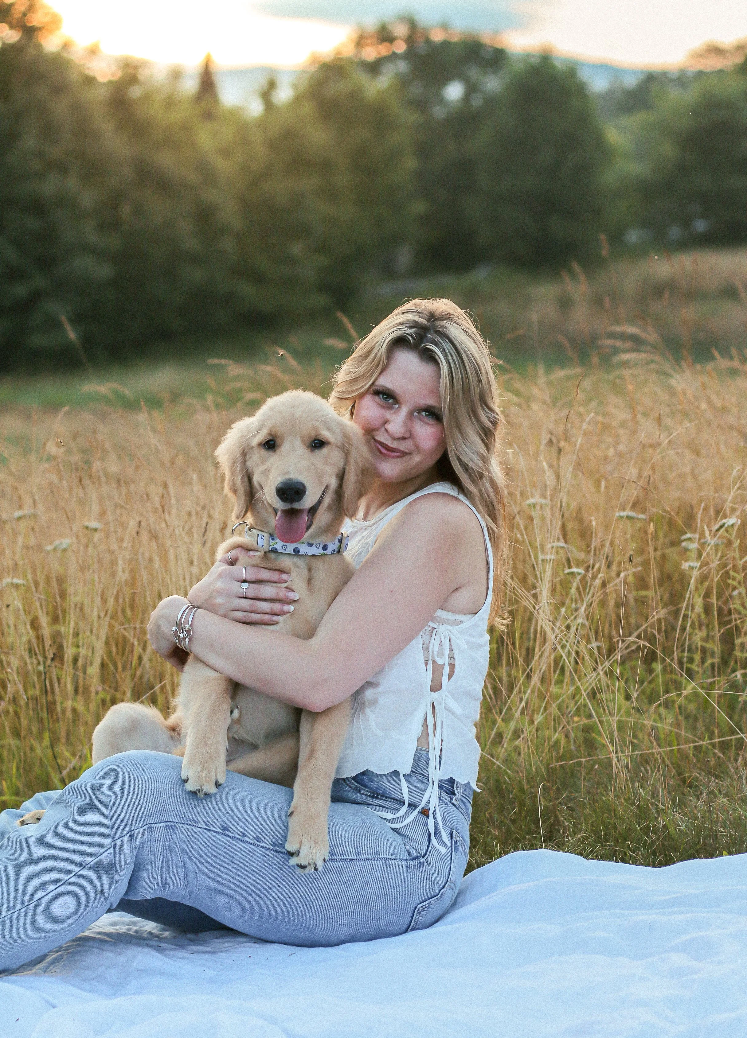 A woman sitting on a blanket in a field holding a yellow retriever puppy, with trees and a sunset in the background.