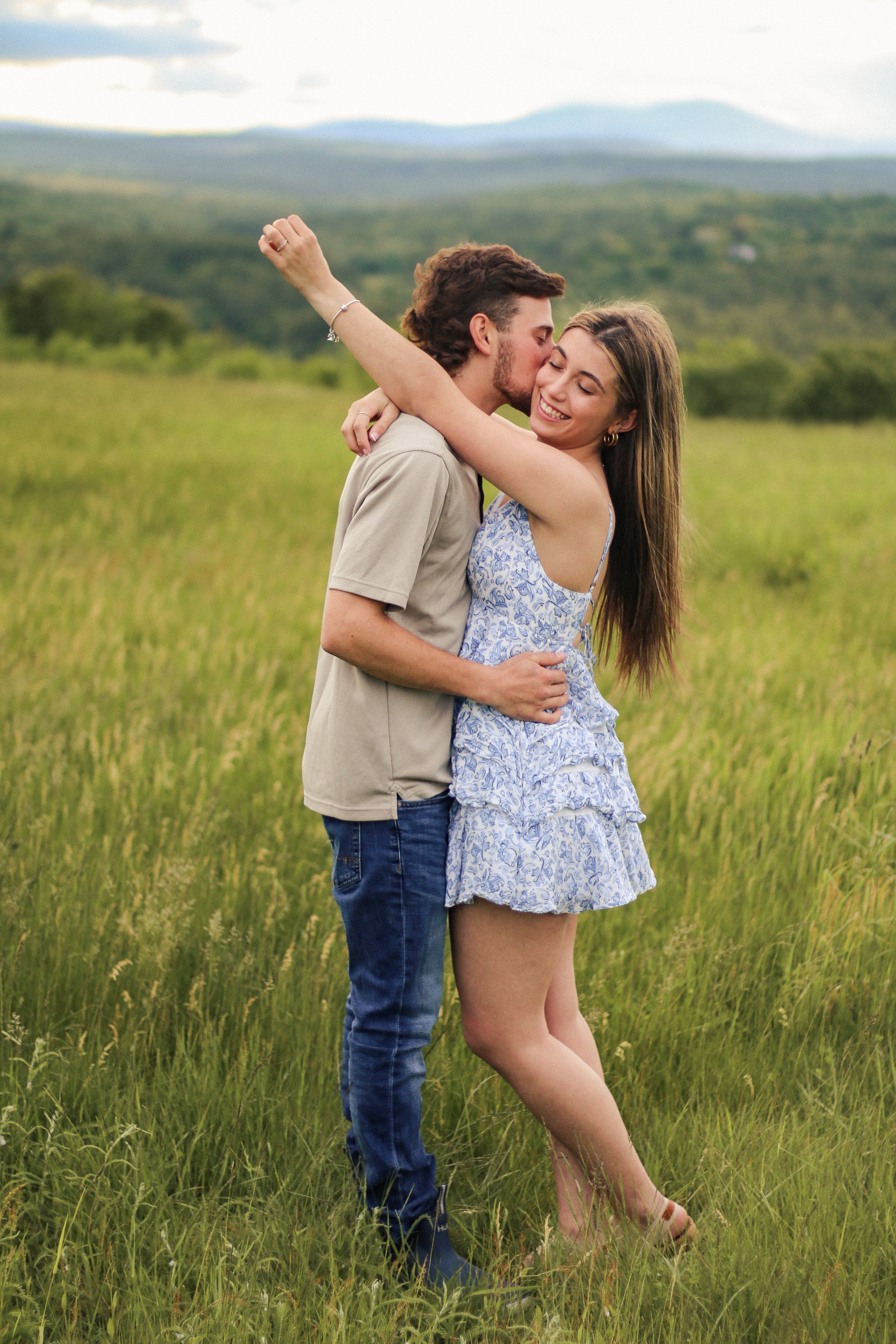 A young couple embracing in a grassy field with mountains in the background, smiling and looking happy.
