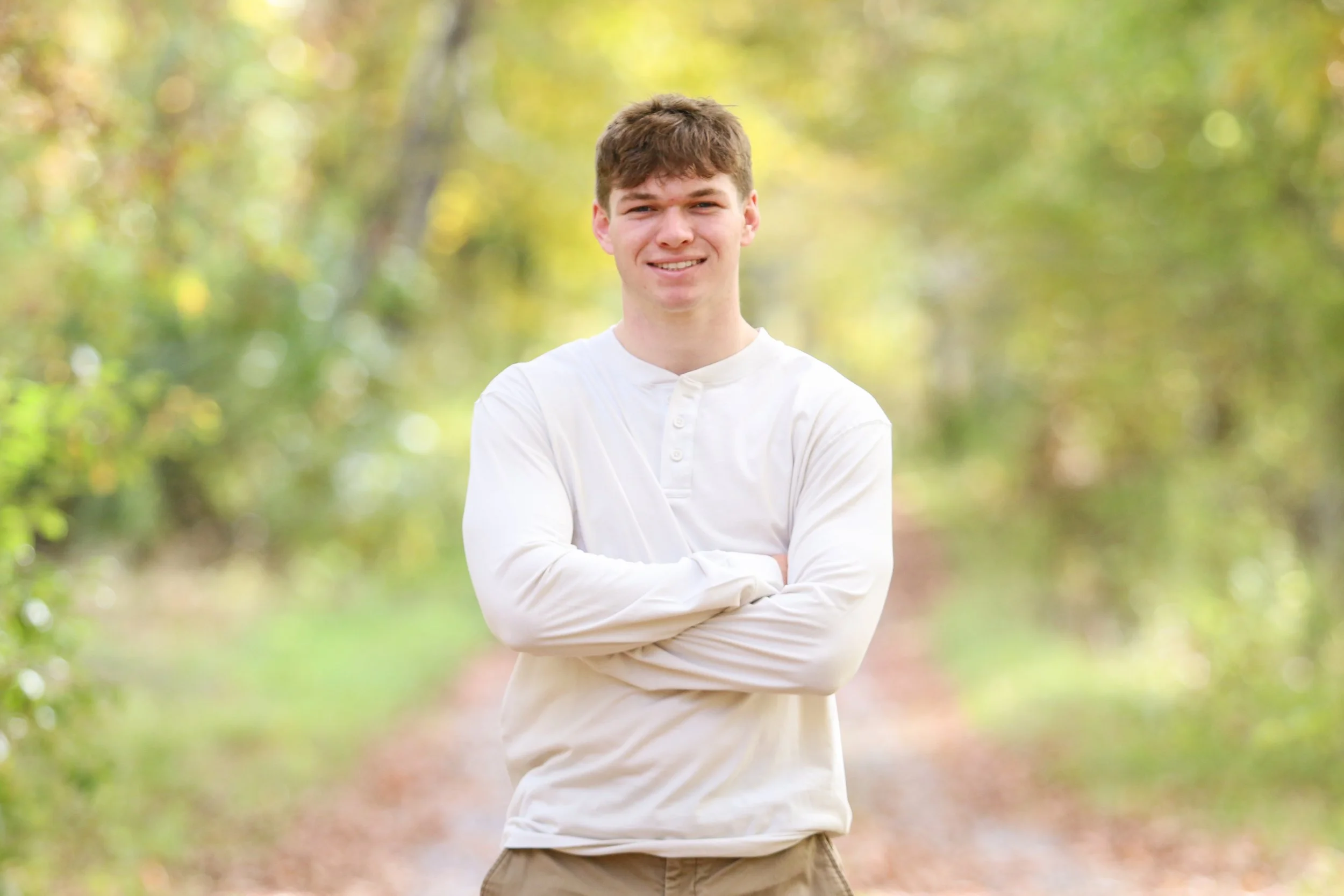 A young man with short brown hair, wearing a white long-sleeve shirt and beige pants, standing with arms crossed on a forest trail, smiling at the camera.