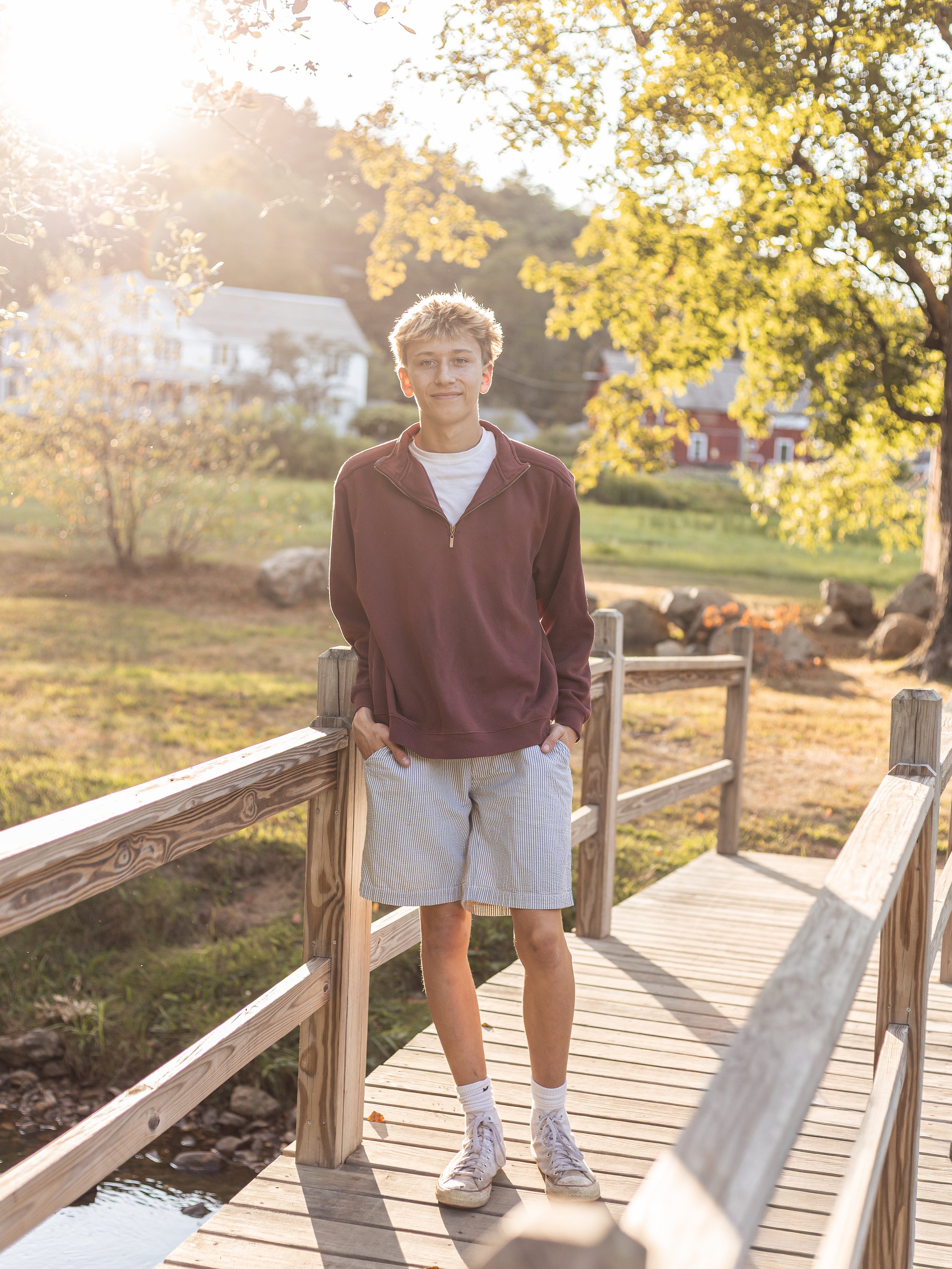 A young man stands on a wooden bridge outdoors during sunset, with trees, grass, rocks, and houses visible in the background.