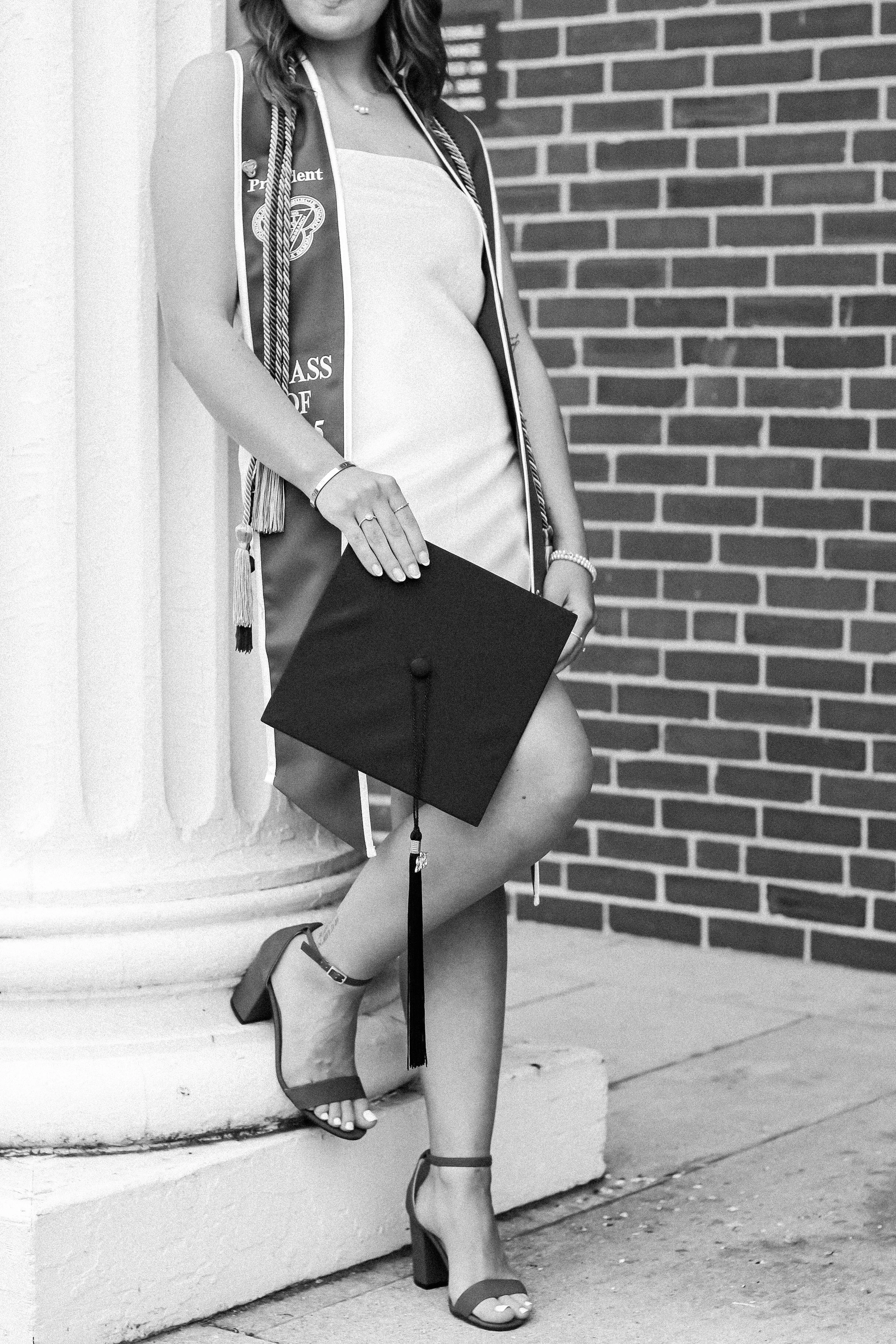 A woman dressed in graduation attire, holding a cap, standing outdoors against a brick wall, with her right leg raised onto a ledge, wearing high heels.