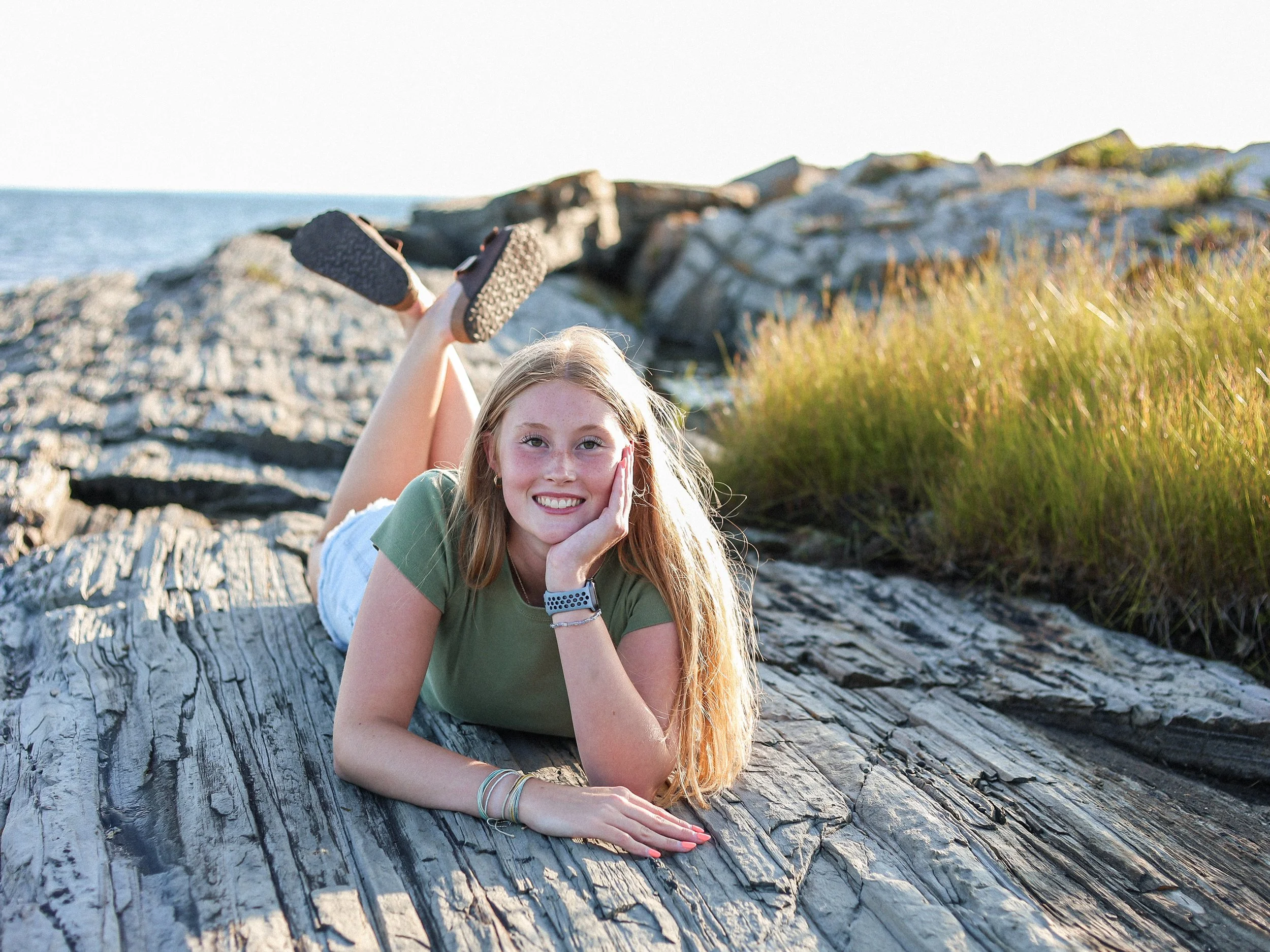 Young woman lying on a large tree trunk near the ocean, smiling at the camera, with rocky shoreline and grass in the background.