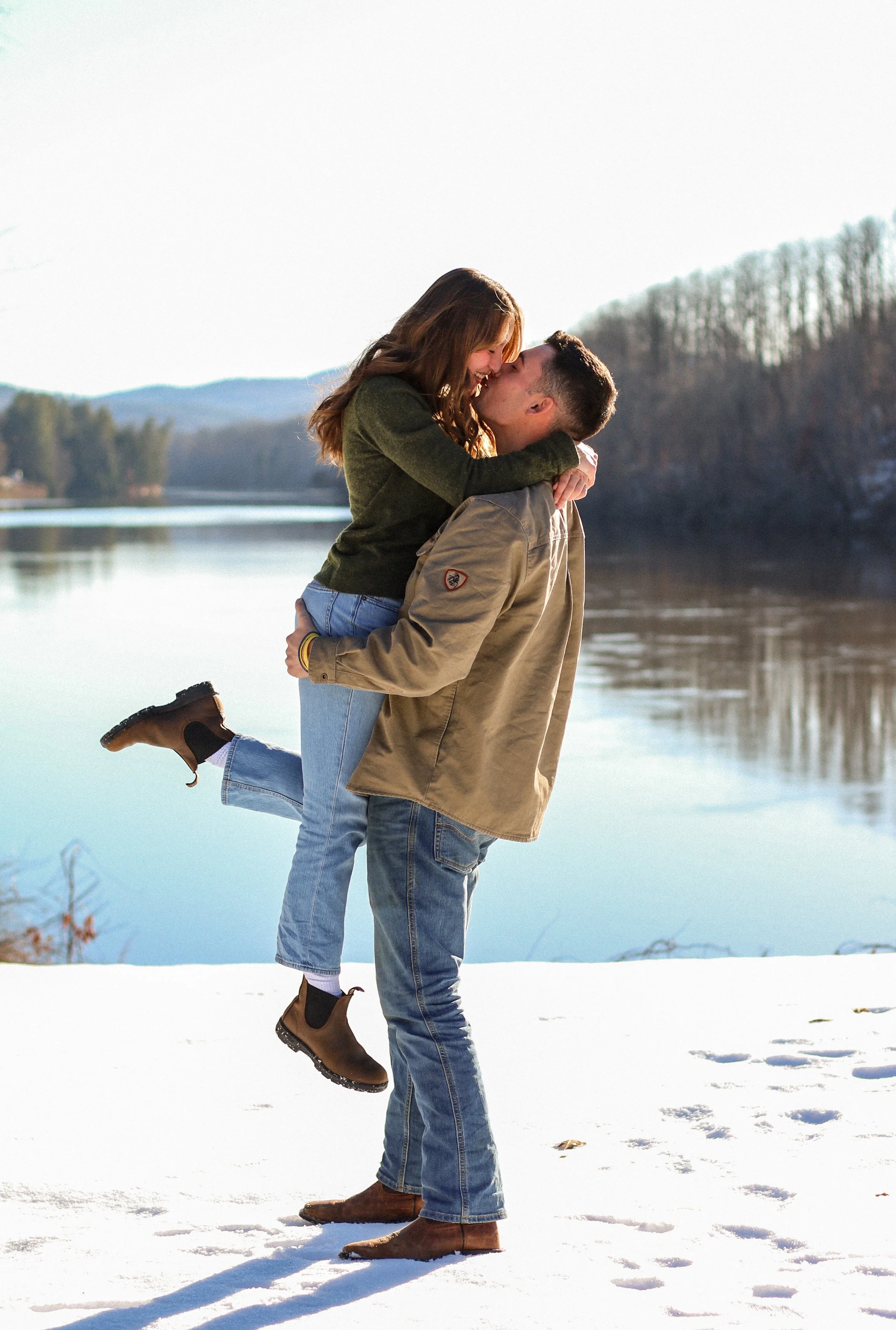 A couple is outdoors in winter, with the man lifting the woman in a joyful embrace by a snowy river, with bare trees and hills in the background.