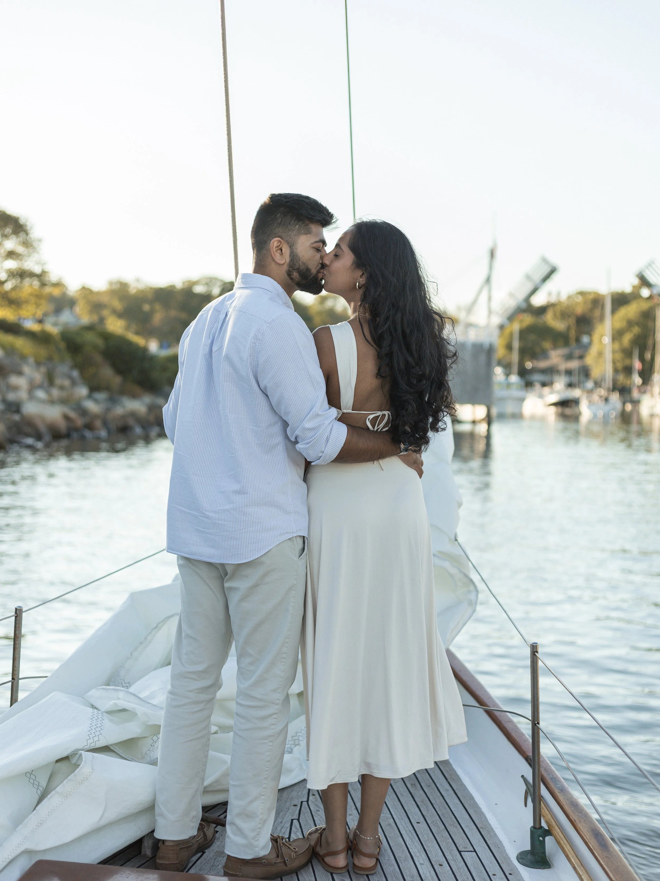 A couple kissing on a boat dock, waterfront with sailboats and trees in the background during sunset.