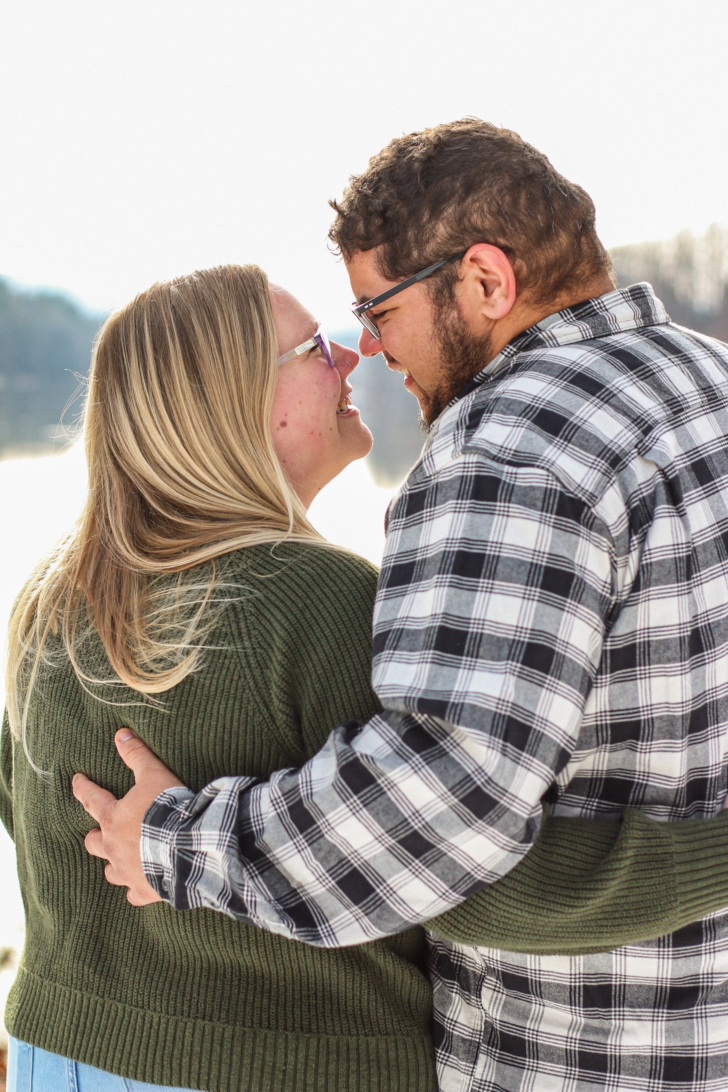 A young couple smiling and touching foreheads outdoors near a body of water on a bright day.