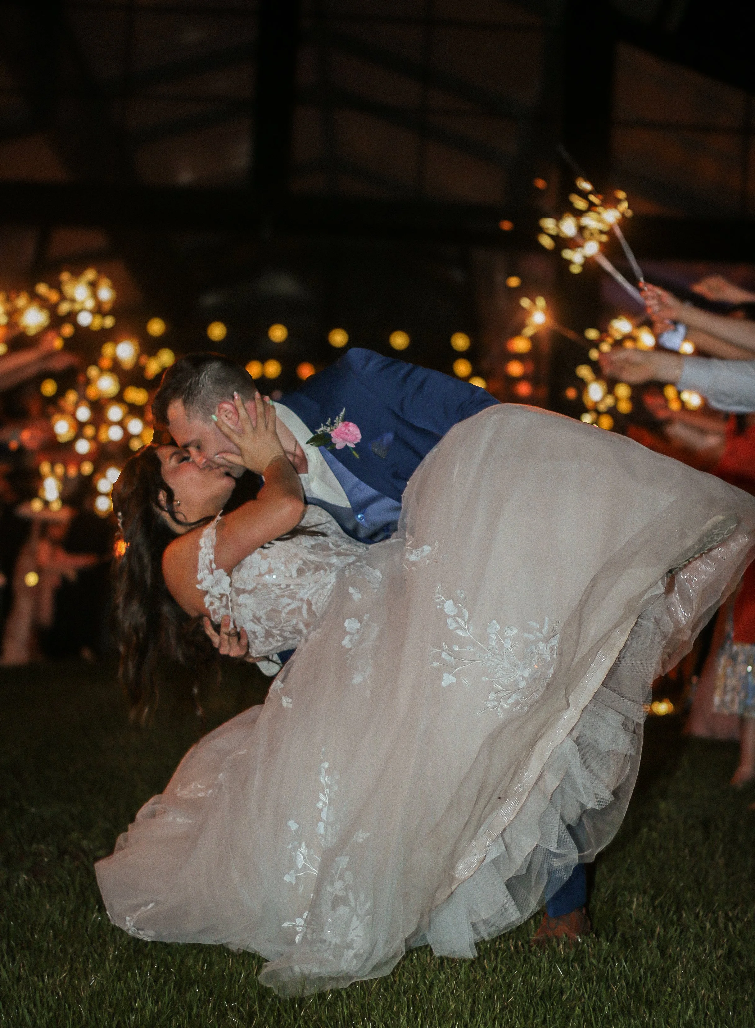 A bride and groom kiss during their wedding reception, with the groom dipping the bride as guests hold sparklers in the background.
