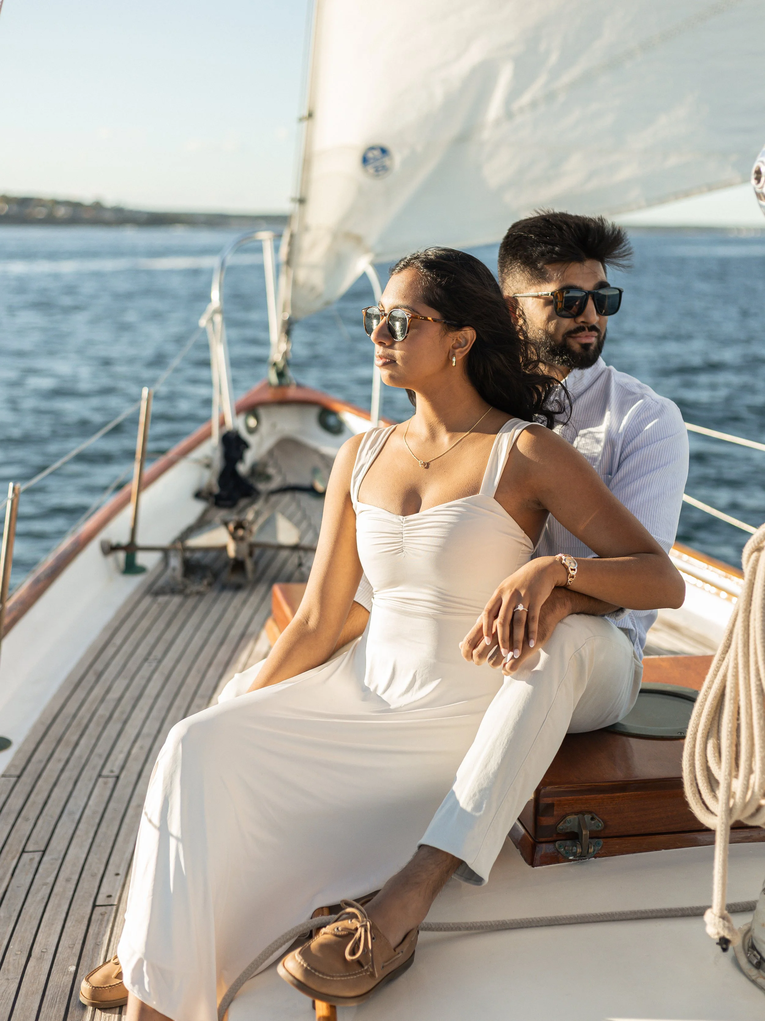 A couple sitting on a sailboat, enjoying a sunny day on the water, with the woman in a white dress and the man in a white shirt and khakis, both wearing sunglasses.