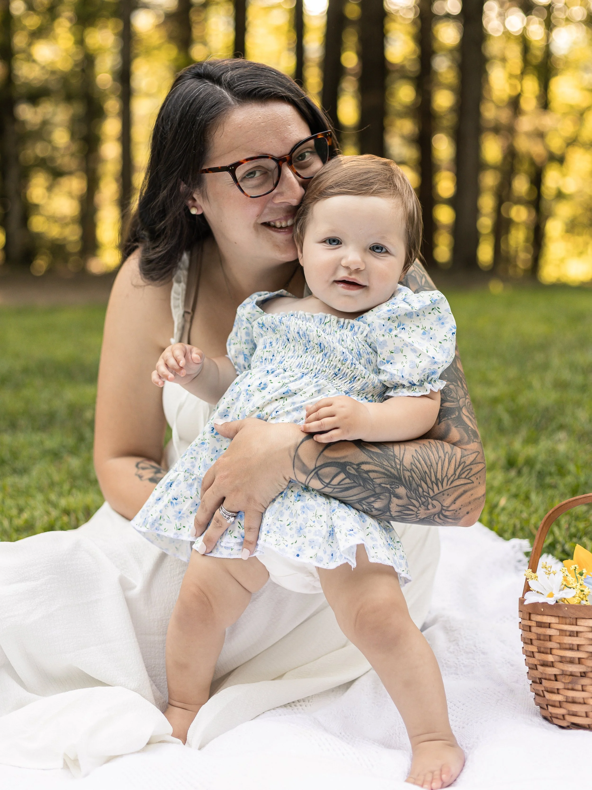 A woman with dark hair, glasses, and tattoos on her arm is sitting on a white blanket outdoors, holding a young girl in a floral dress. The girl has light brown hair and blue eyes. They are in a forest with sunlight filtering through the trees, and t