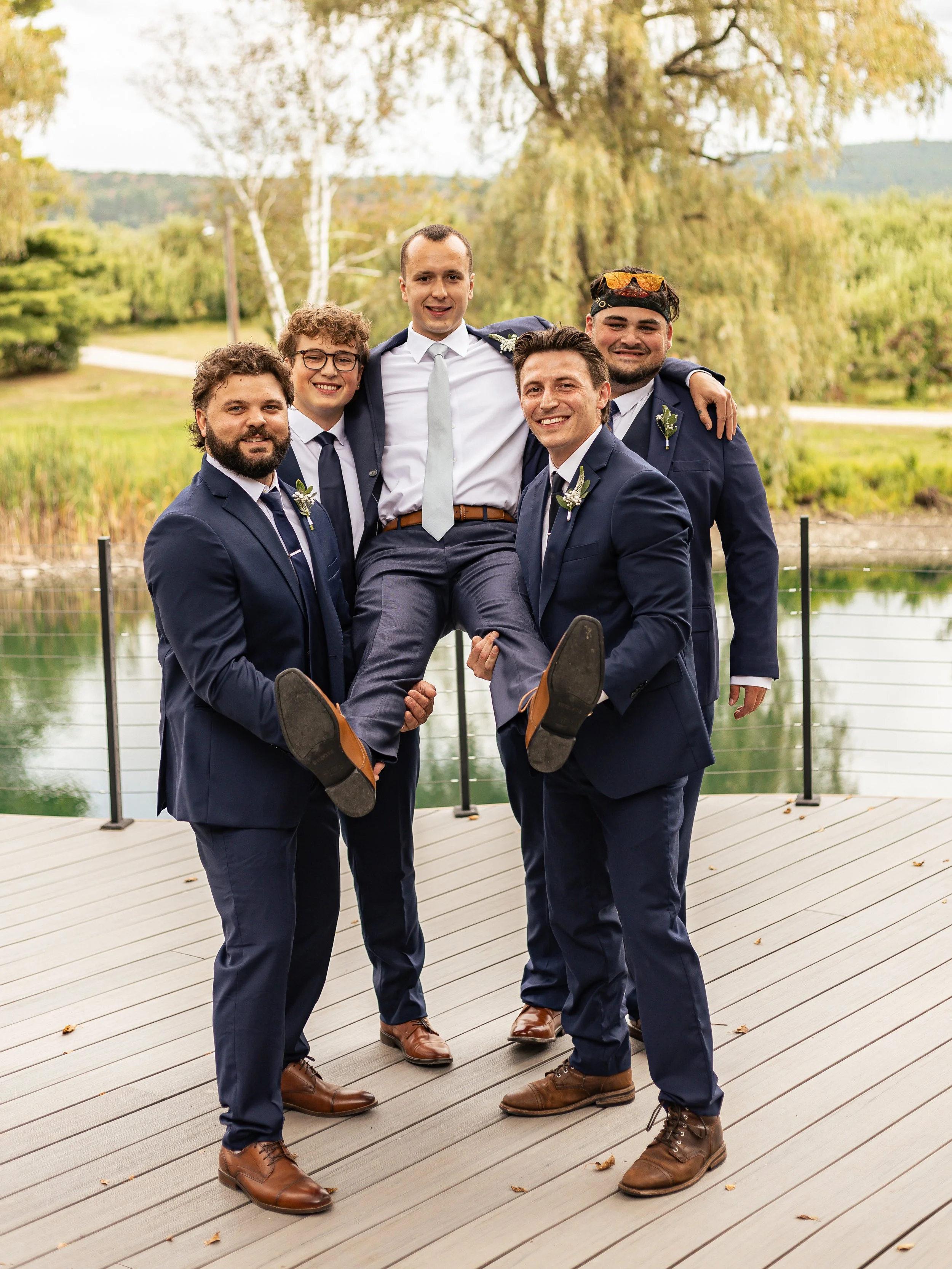 Group of five men in suits, two with boutonnières, lifting a man in a white shirt and tie with sunglasses on his head, all smiling and standing outdoors near a pond with trees in the background.