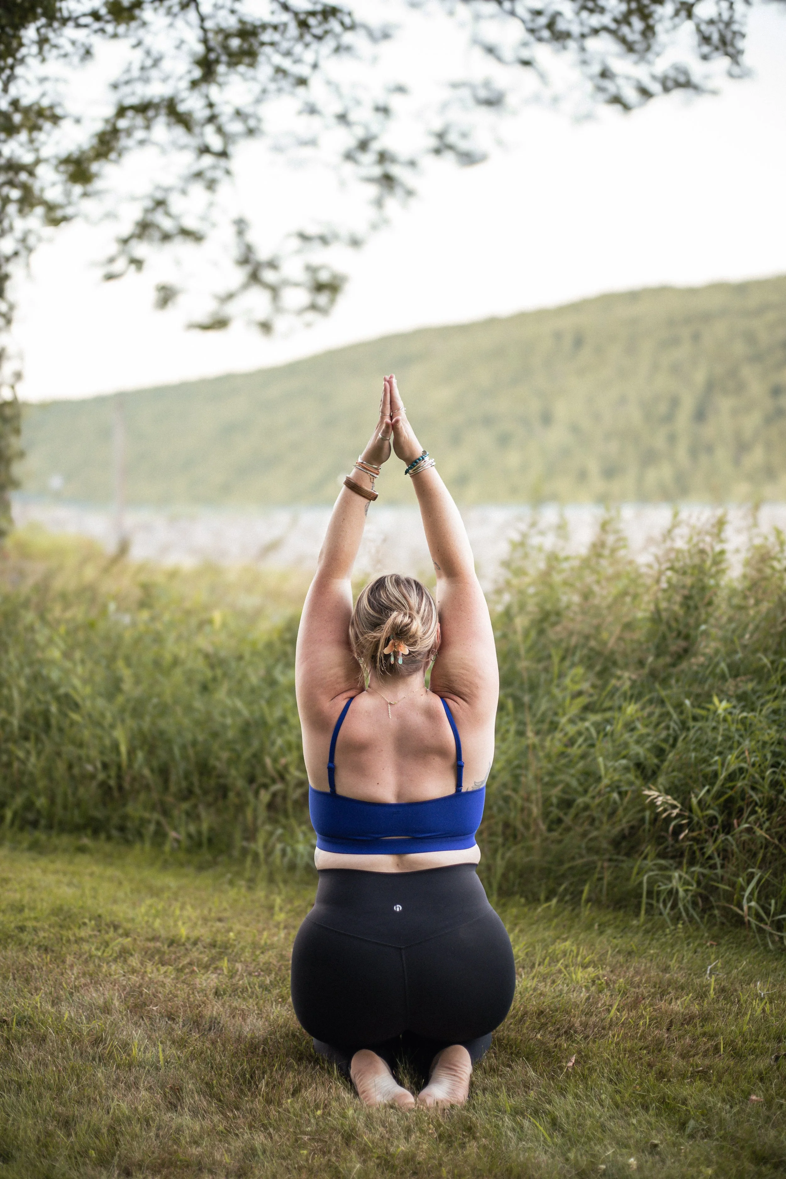 A woman practicing yoga outdoors, kneeling on grass with arms raised and hands together in a prayer position, facing away from the camera in a natural setting with trees and hills in the background.
