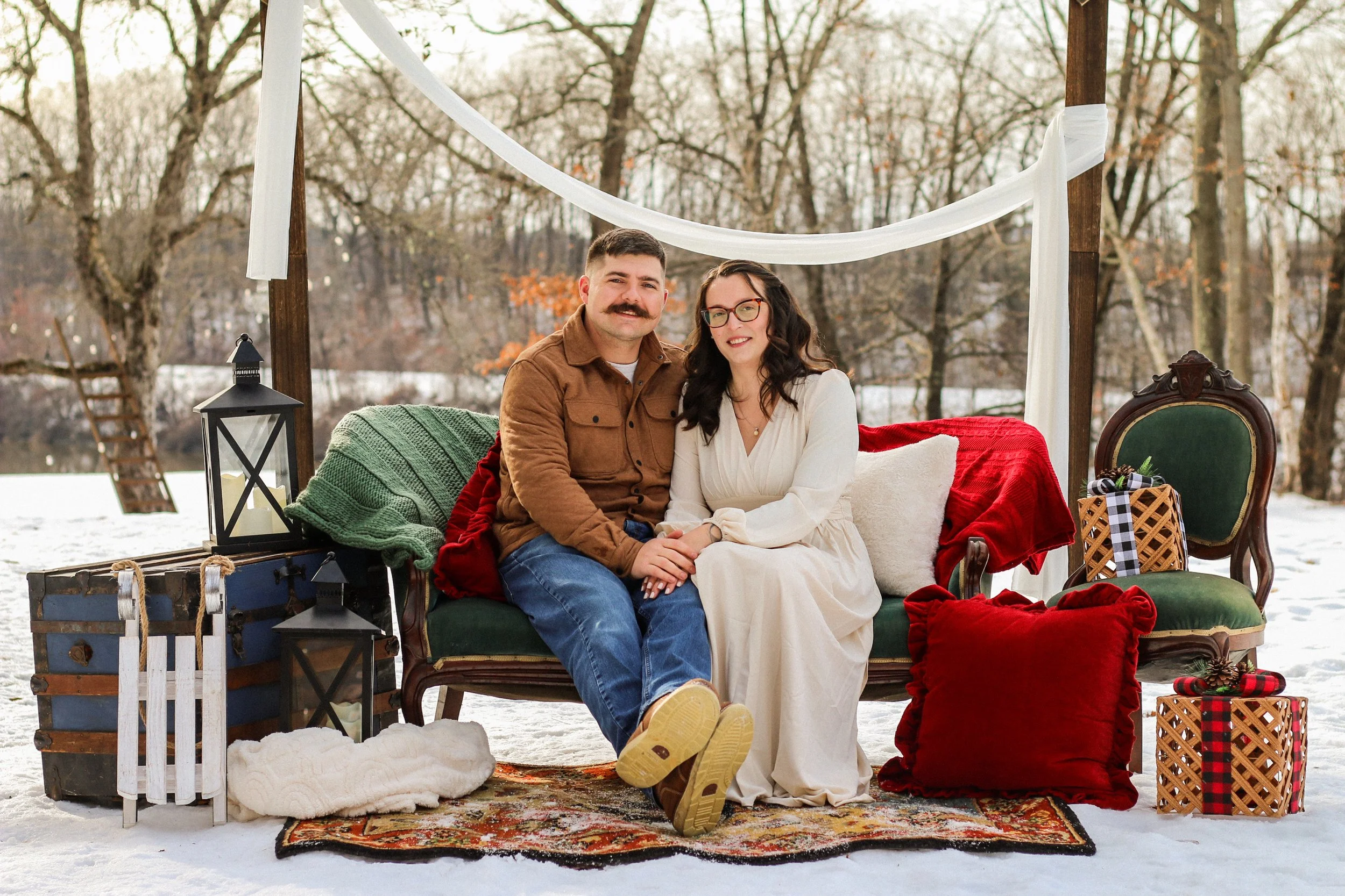 A couple sitting on a vintage sofa in a snowy outdoor setting decorated for Christmas, with wrapped gifts, pillows, lanterns, and a colorful rug.