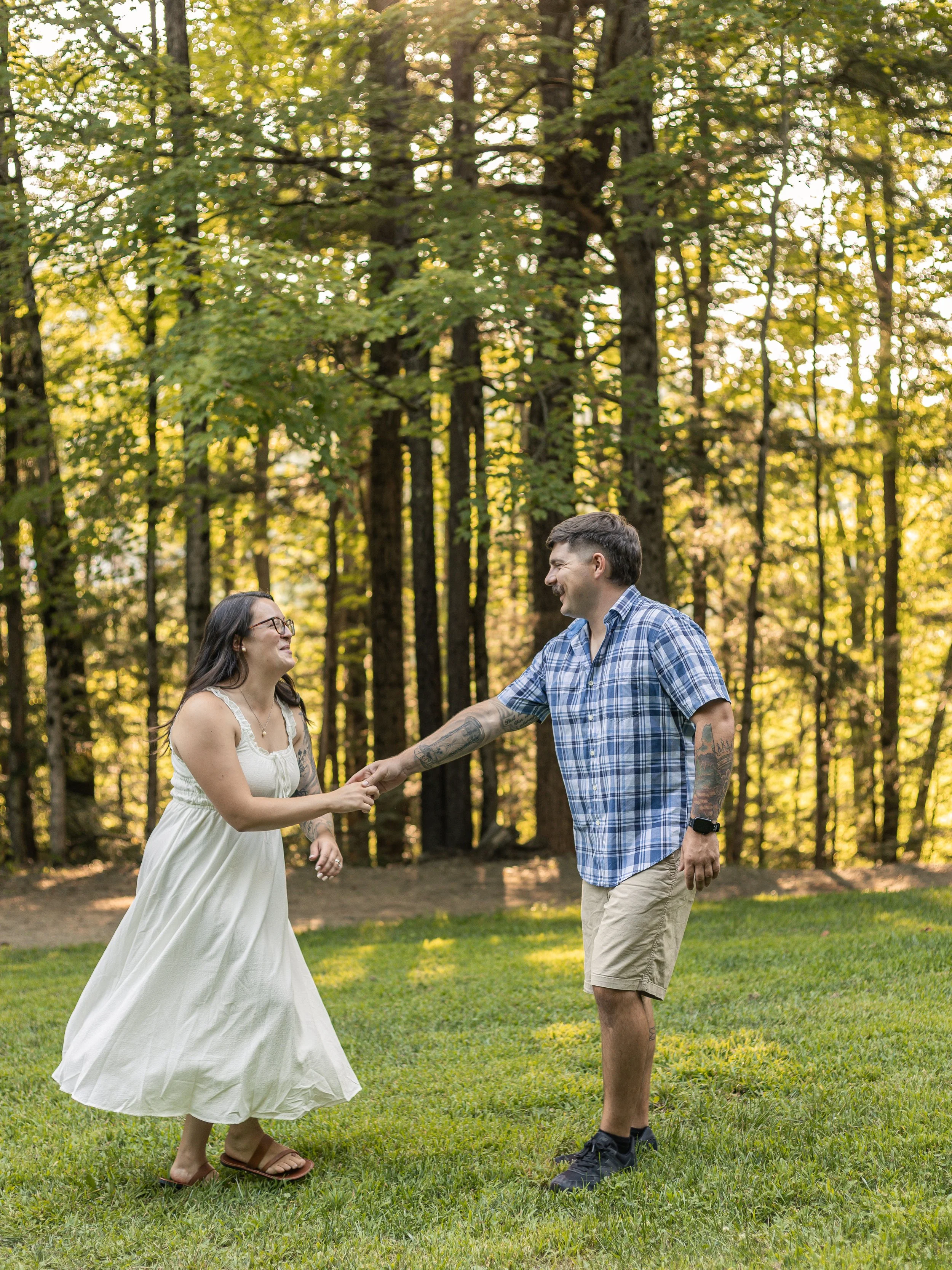 A couple holding hands and smiling in a wooded park during daylight, with green grass and tall trees in the background.