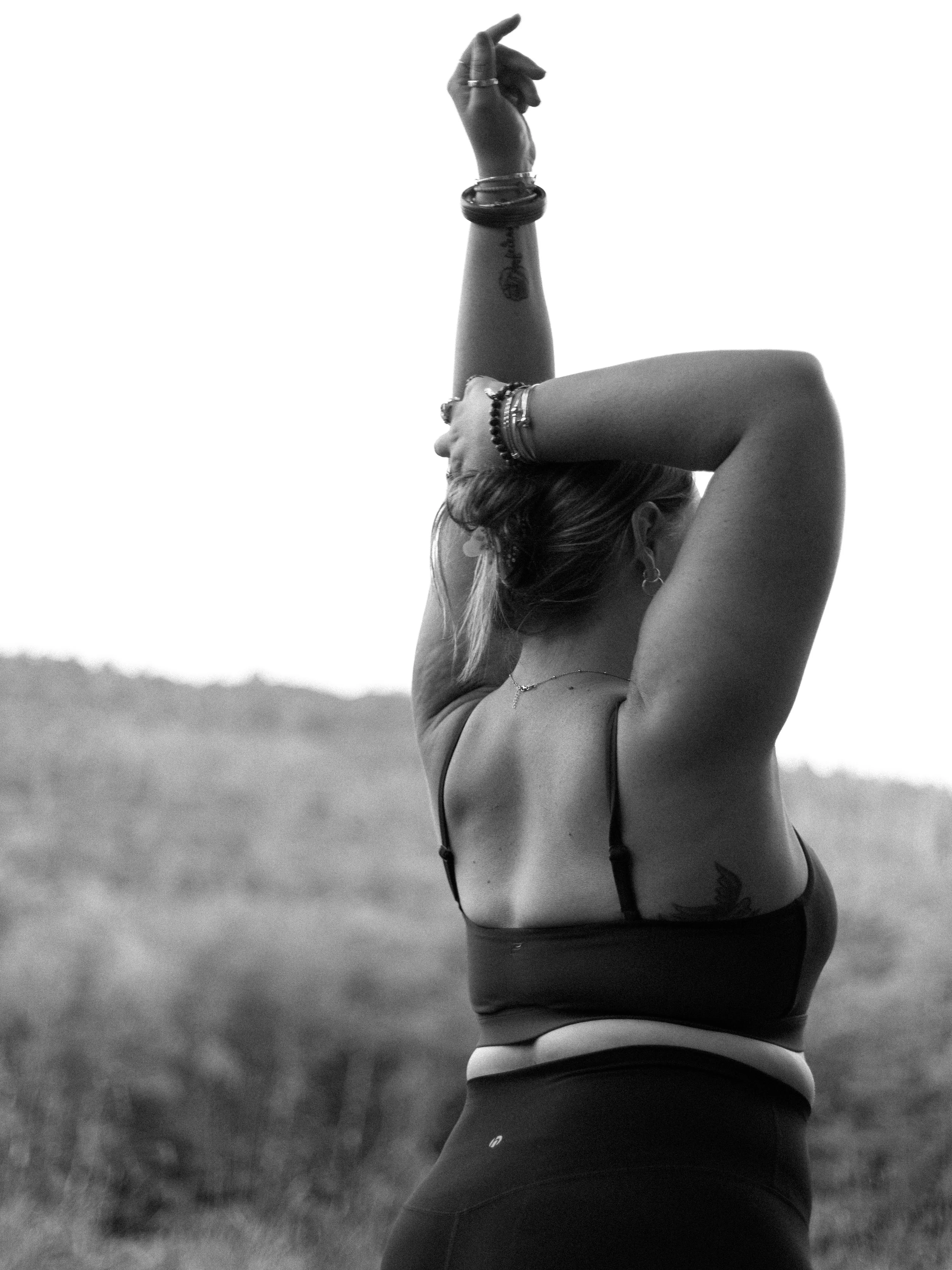 A woman practicing yoga outdoors, stretching with one arm raised above her head and the other bent behind her neck, with a natural landscape in the background.