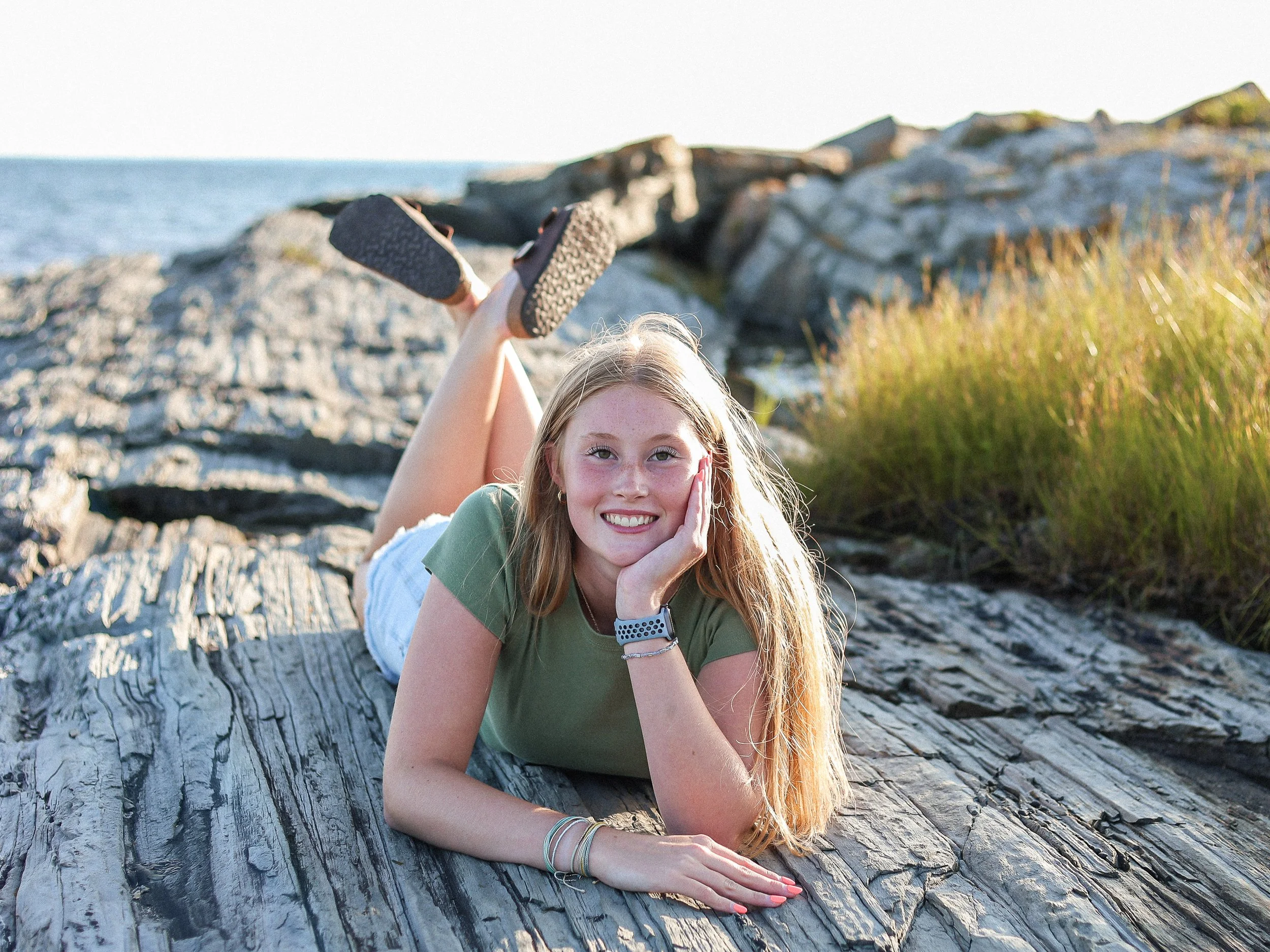 A young woman with long blonde hair lying on a large piece of driftwood near the ocean, smiling at the camera, with grassy rocks and water in the background.