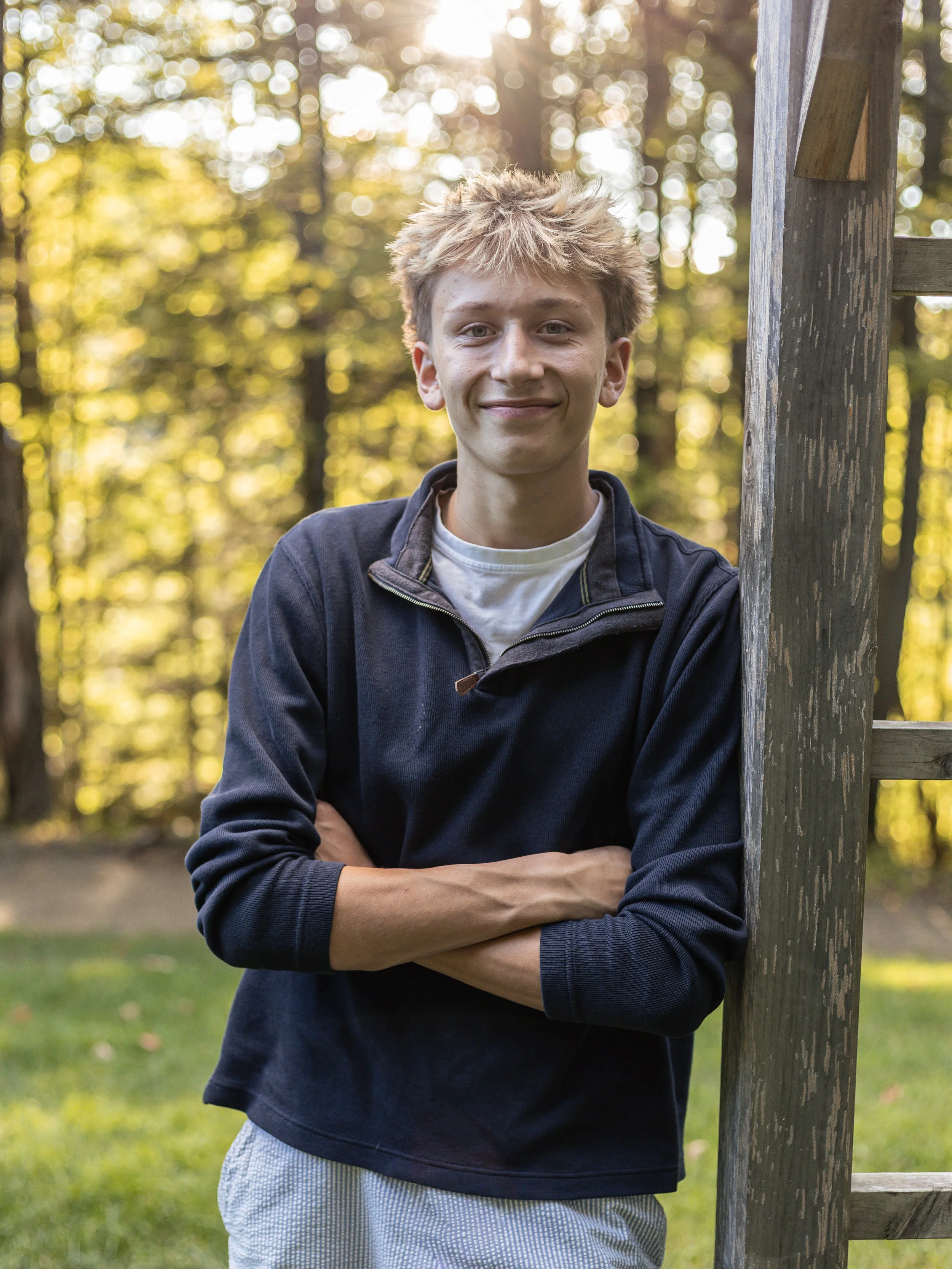 A young man with blond, spiky hair, wearing a navy zip-up jacket over a white shirt, standing outdoors with trees and sunlight in the background, leaning against a wooden post with his arms crossed and smiling.