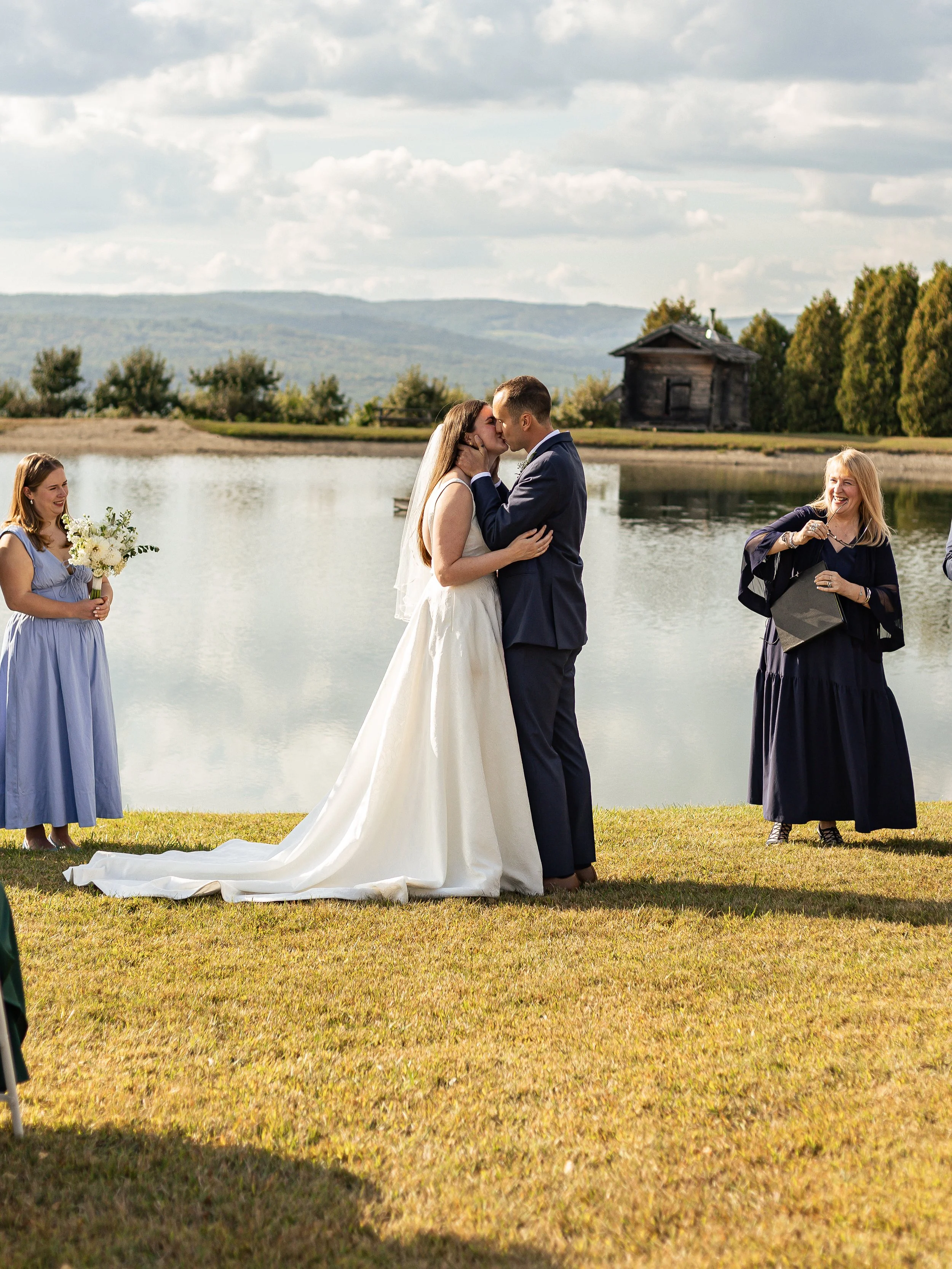 A couple getting married outdoors by a lake, with the bride in a white dress and long train, and the groom in a navy suit, sharing a kiss. Two women, one holding a bouquet and the other with a purse, stand nearby, smiling under a partly cloudy sky.