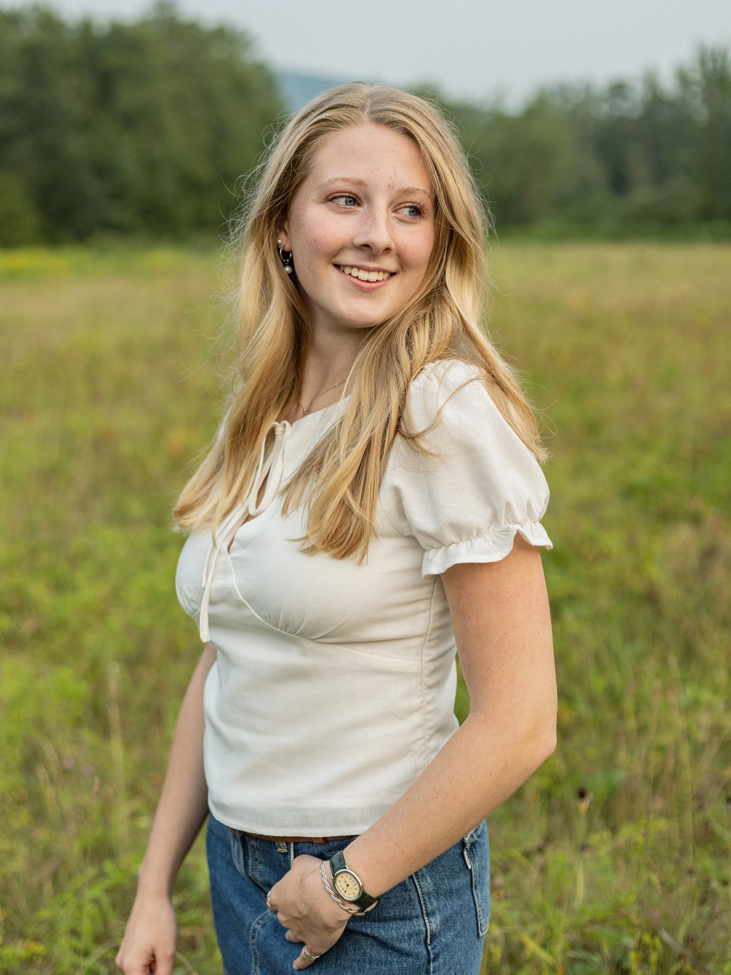 Young woman with long blonde hair smiling outdoors in a field, wearing a white blouse and jeans.
