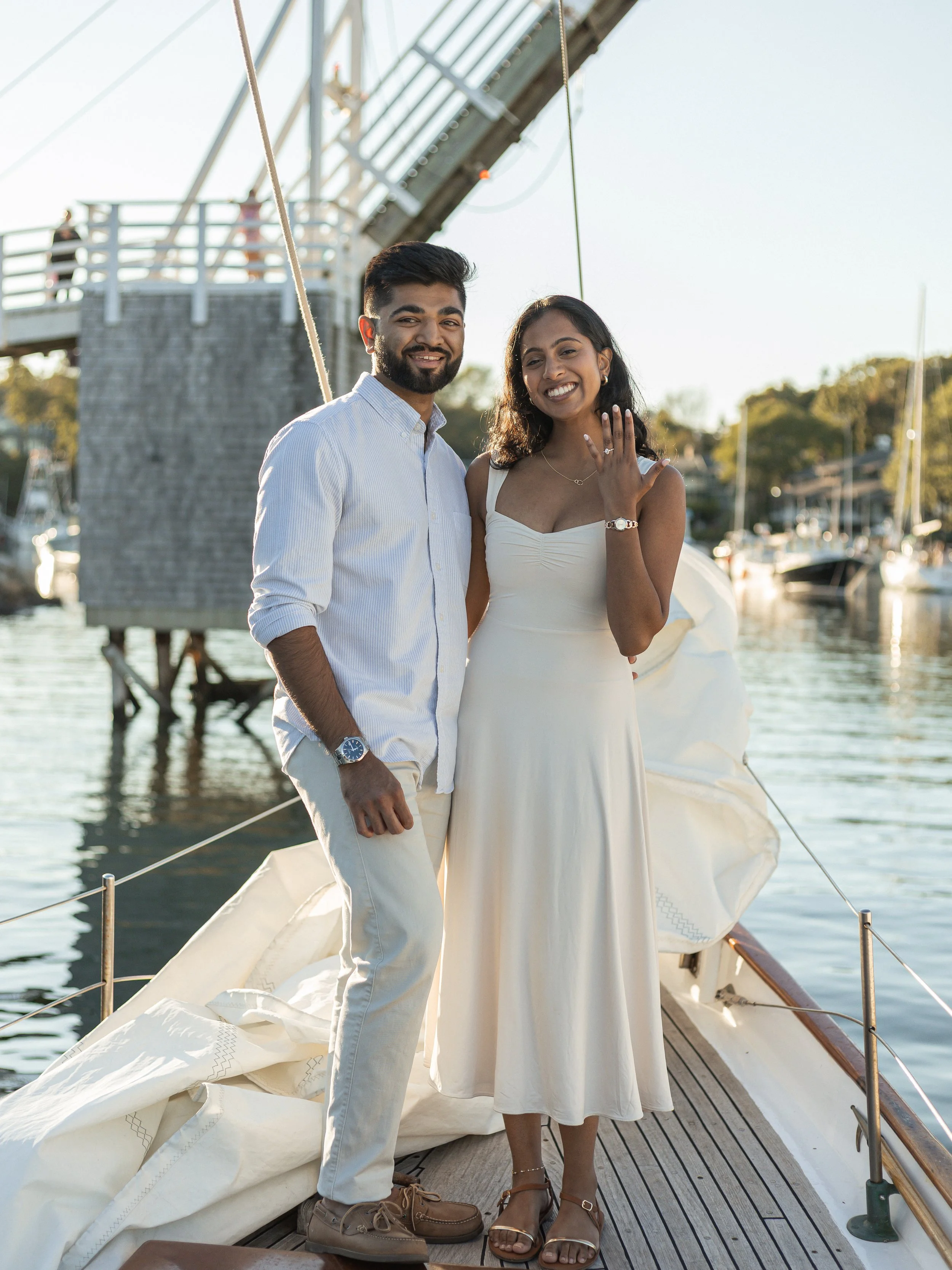 A couple standing on a sailboat dock, with a marina and a wooden bridge in the background, during sunset.
