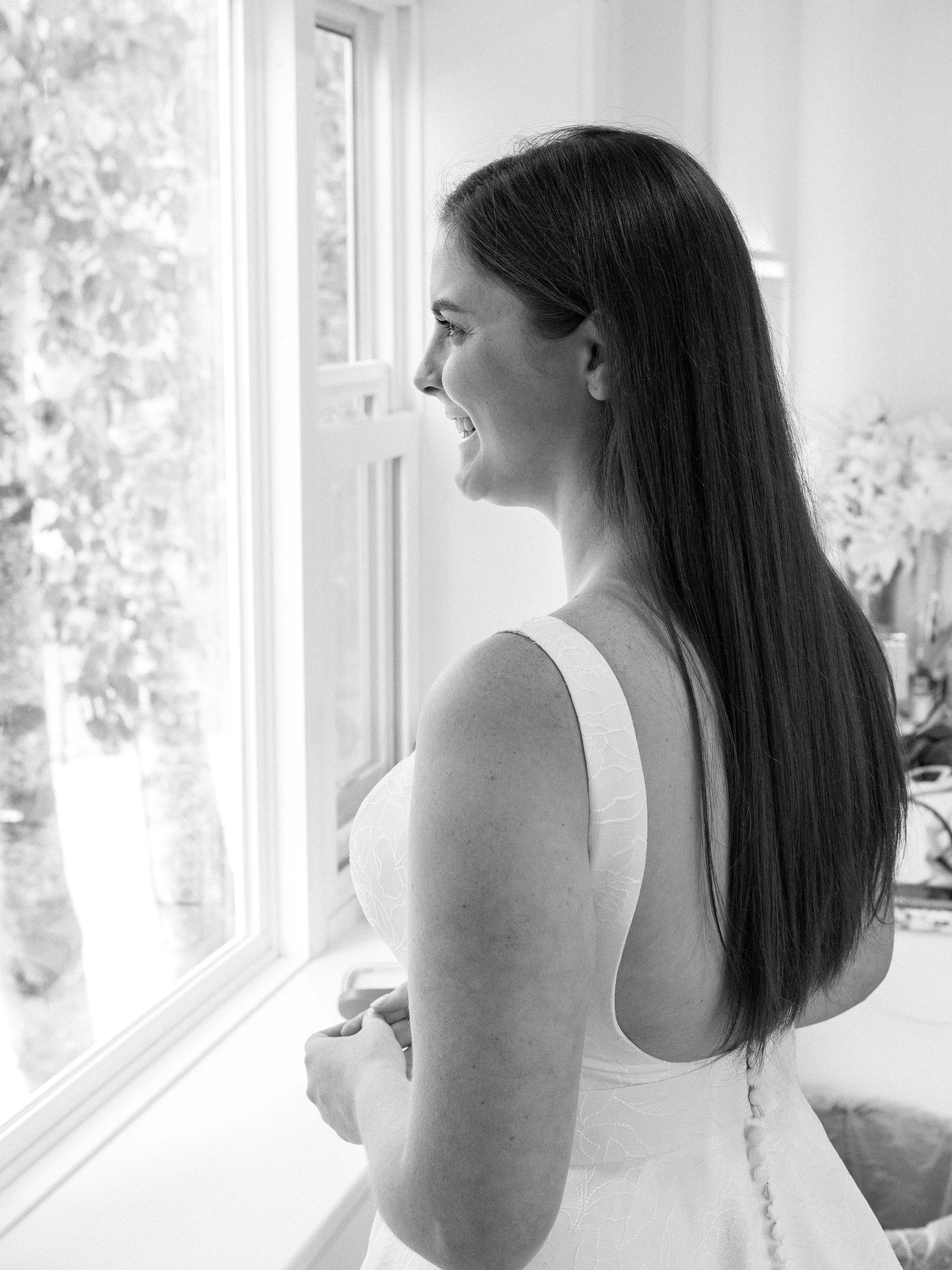 A woman looking out a window, smiling, wearing a sleeveless dress, with a flower arrangement in the background.