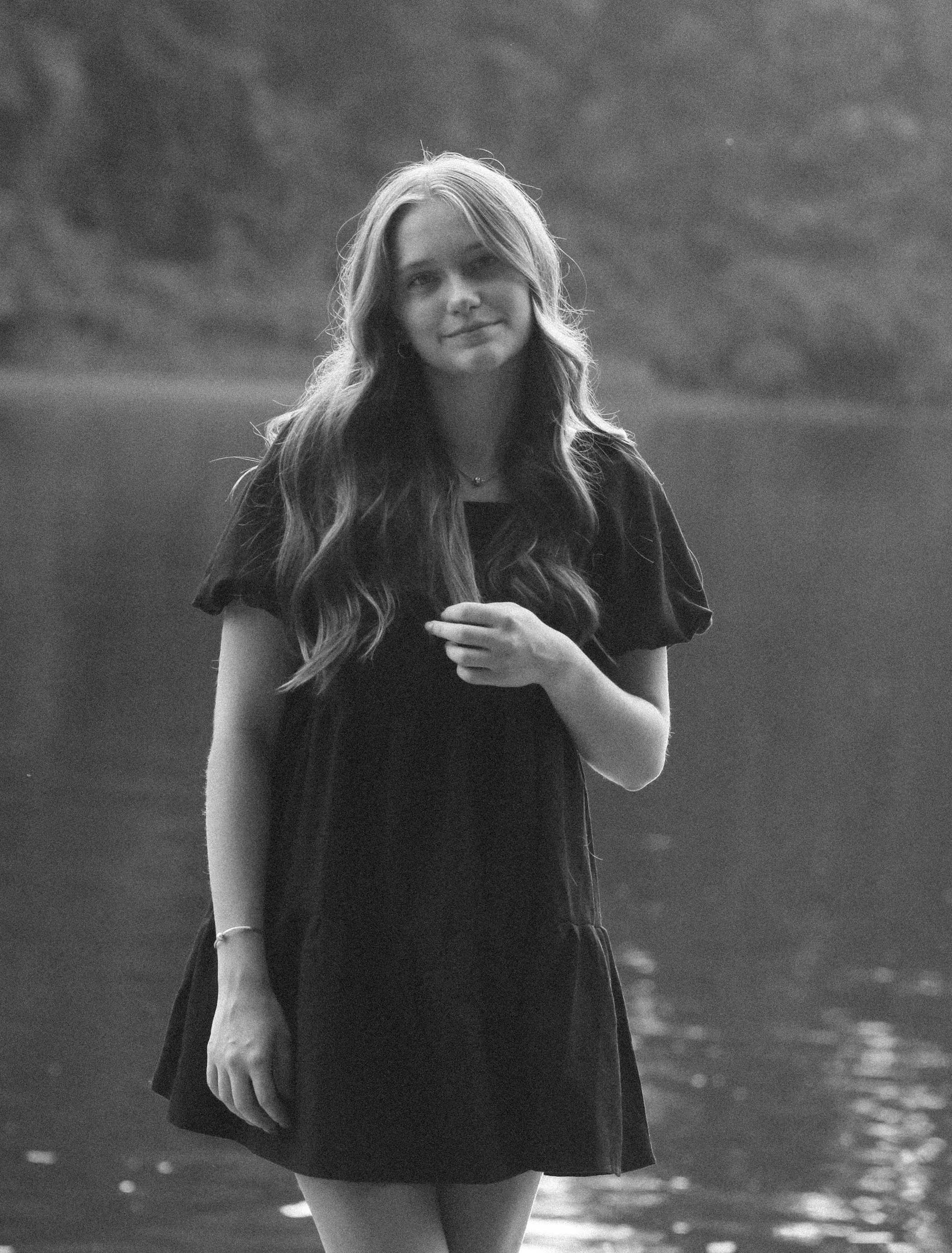A young woman with long, wavy hair stands outdoors near a body of water, wearing a short-sleeved dress and smiling softly.