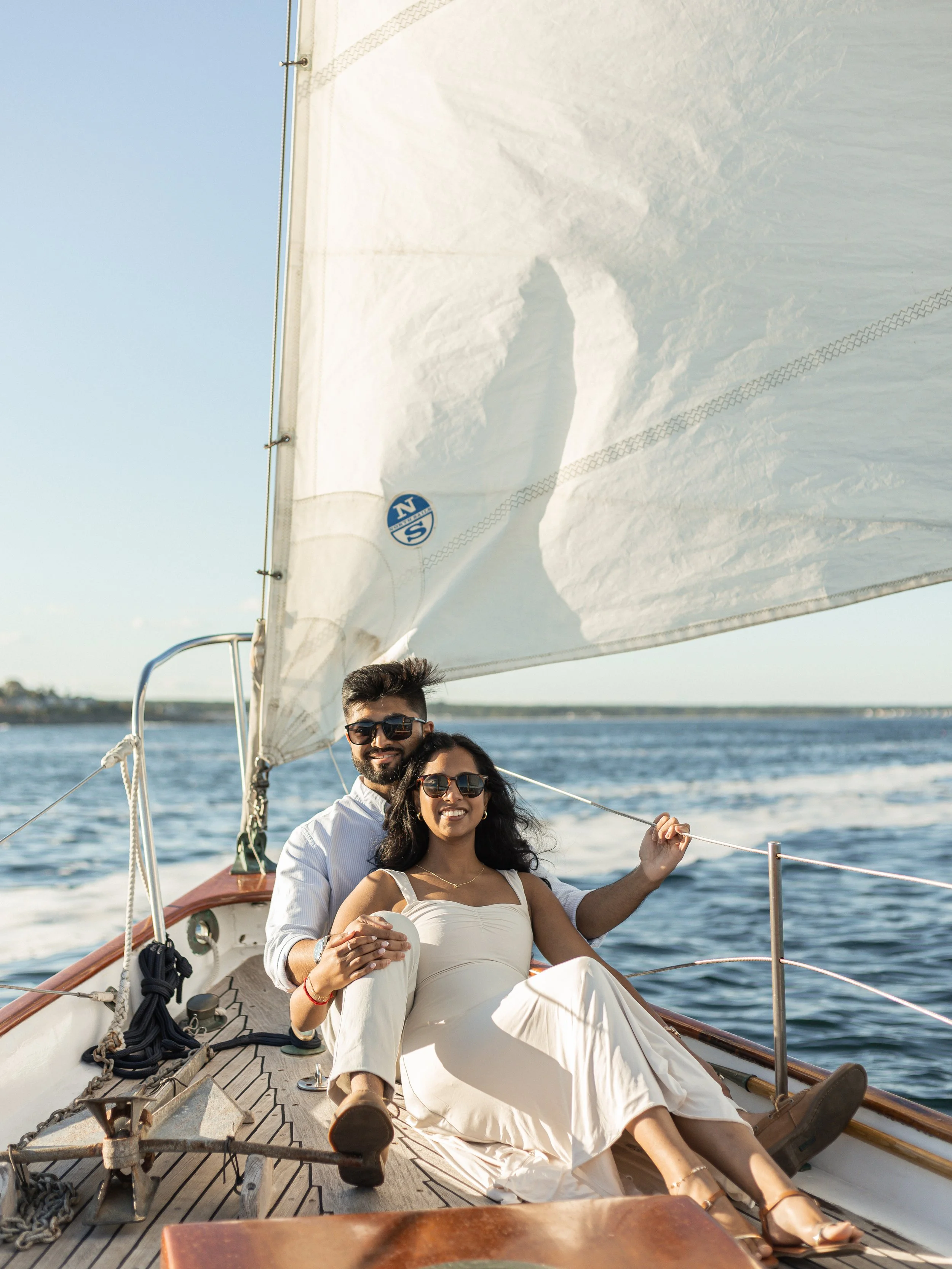 Two people sitting on a sailboat smiling at the camera, with water and a clear sky in the background.